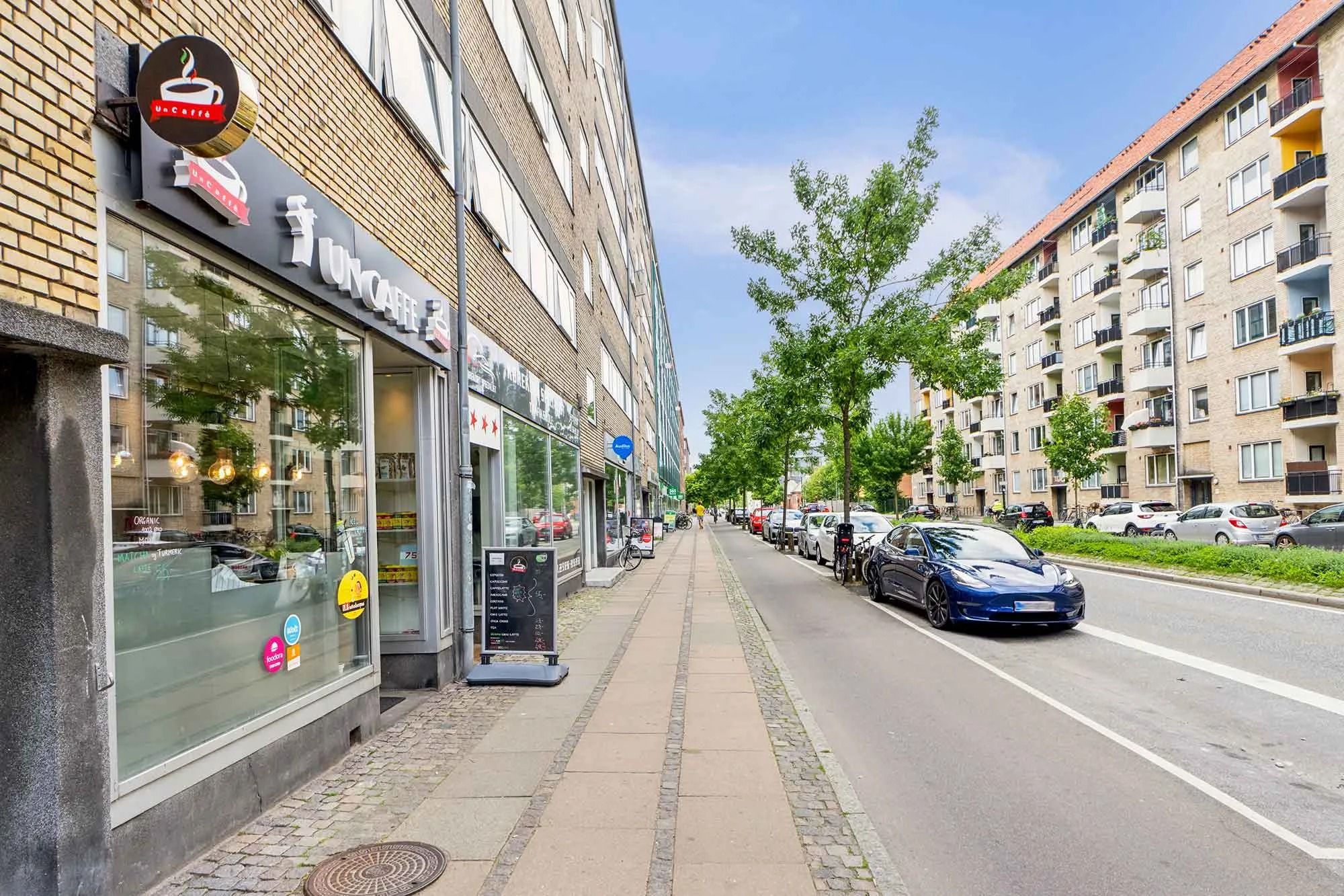 Forretningslokaler med forretningsskilte og Tesla bilparkeret langs en gade med træer og lejlighedsbygninger