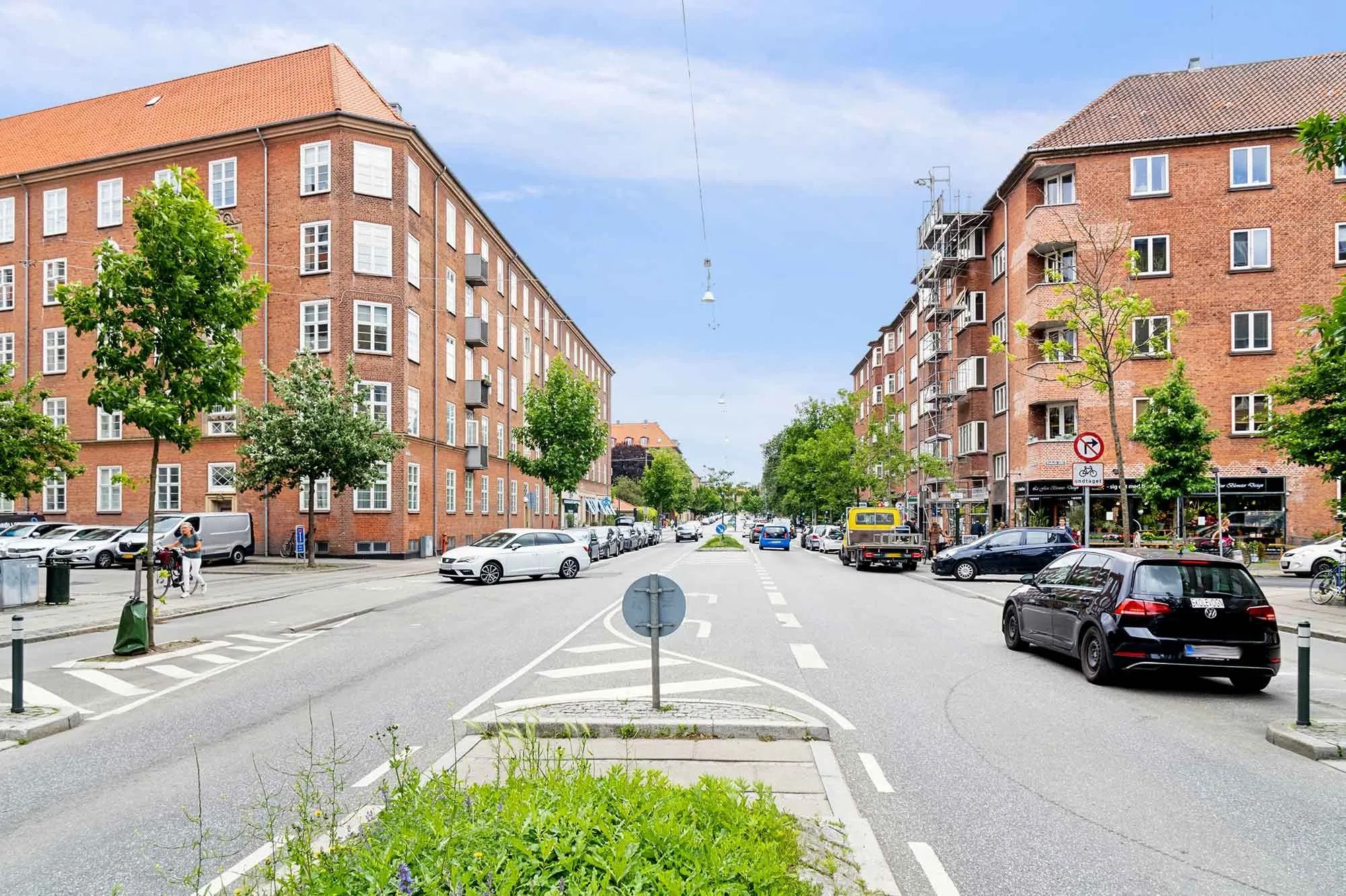 På en bygade med biler og busser er der flere huse og butikker med mange vinduer og nogle skilter med cykelforbud