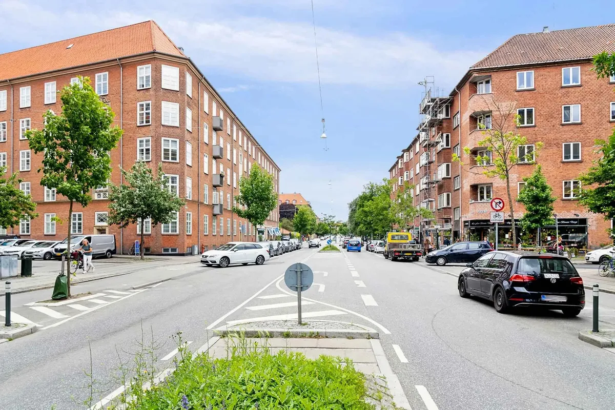 På en bygade med biler og busser er der flere huse og butikker med mange vinduer og nogle skilter med cykelforbud
