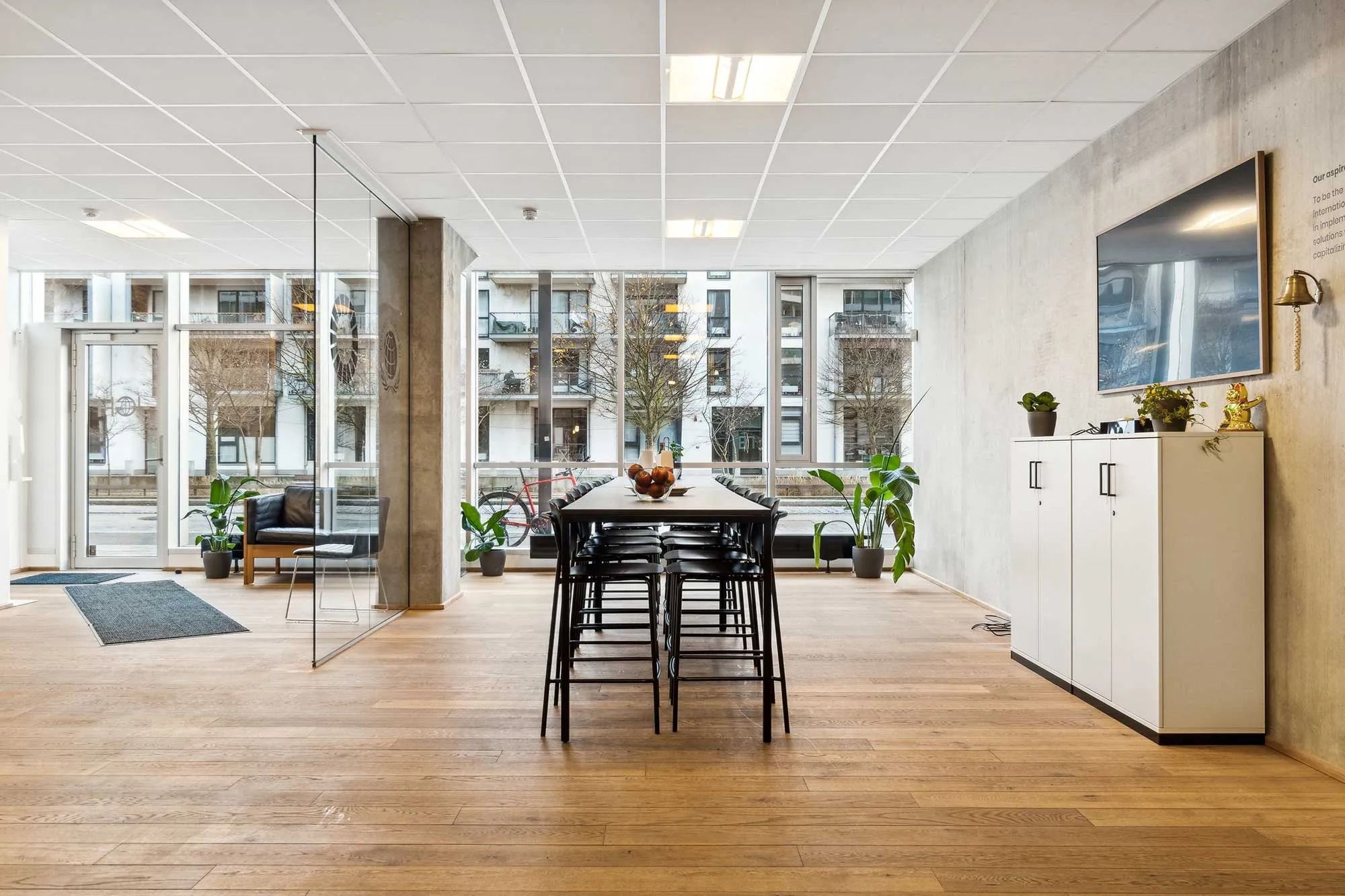 Moderne mødelokale med trægulv, glasvægge, møbler, plantager og en stor tv-skærm på væggen