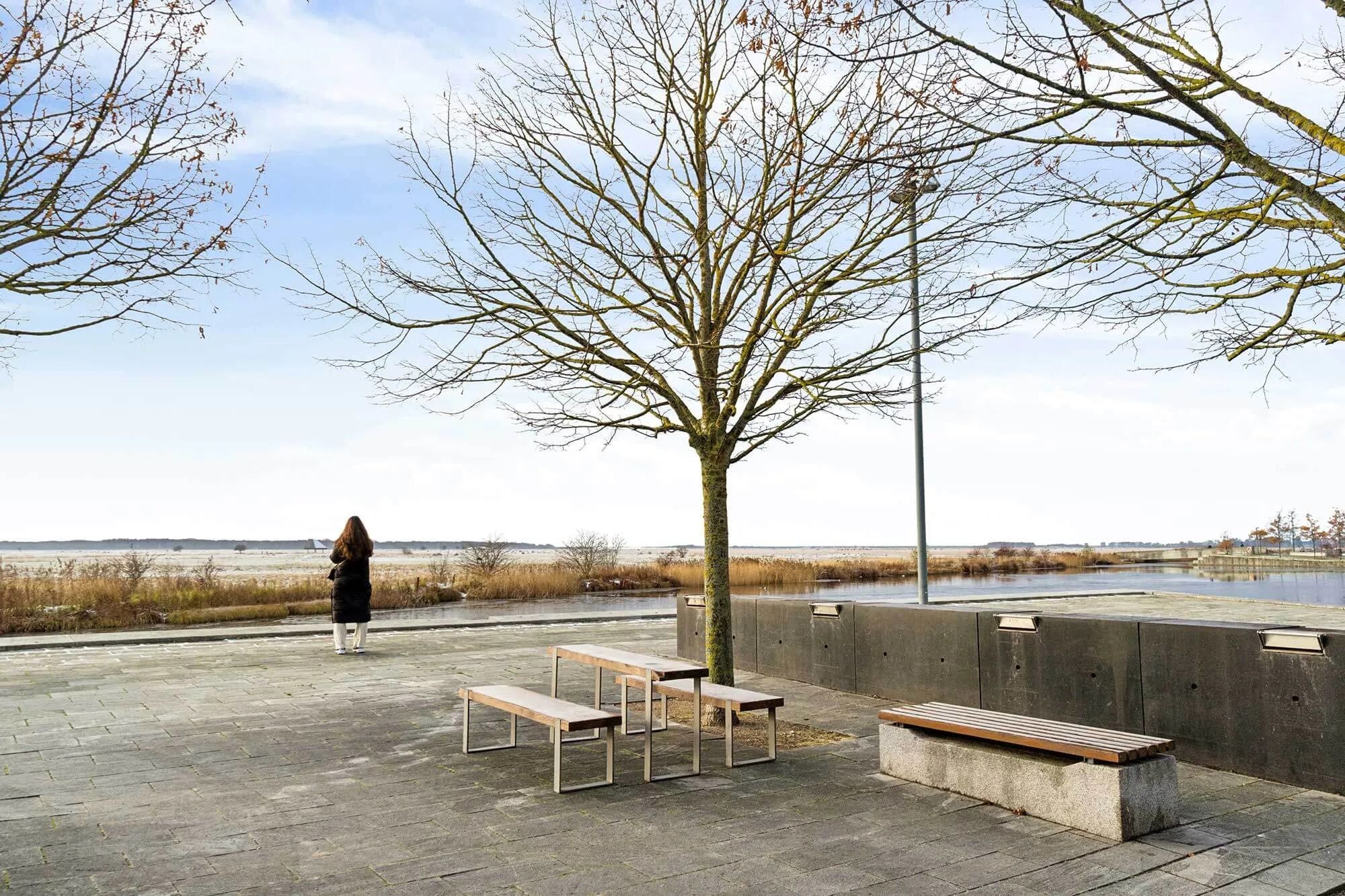 Dame står på promenaden og ser på vandet med bakker og træer i baggrunden