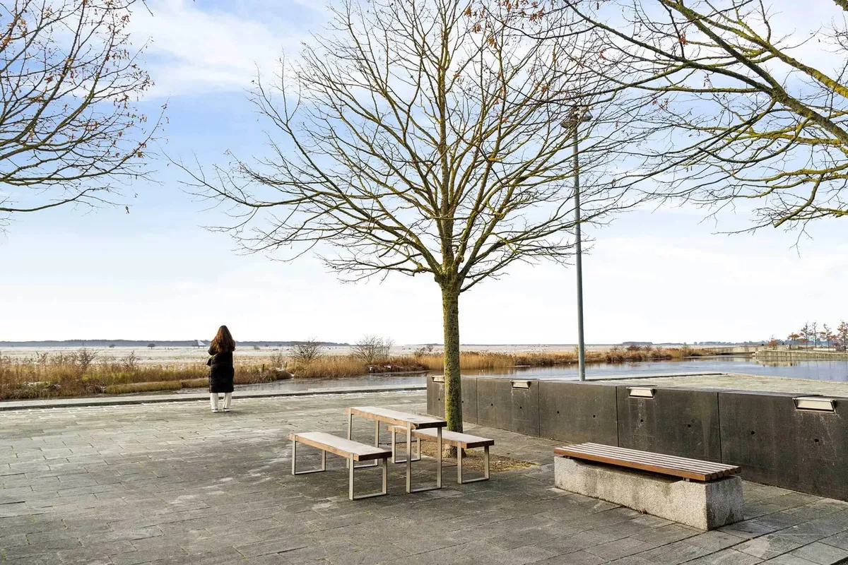 Dame står på promenaden og ser på vandet med bakker og træer i baggrunden
