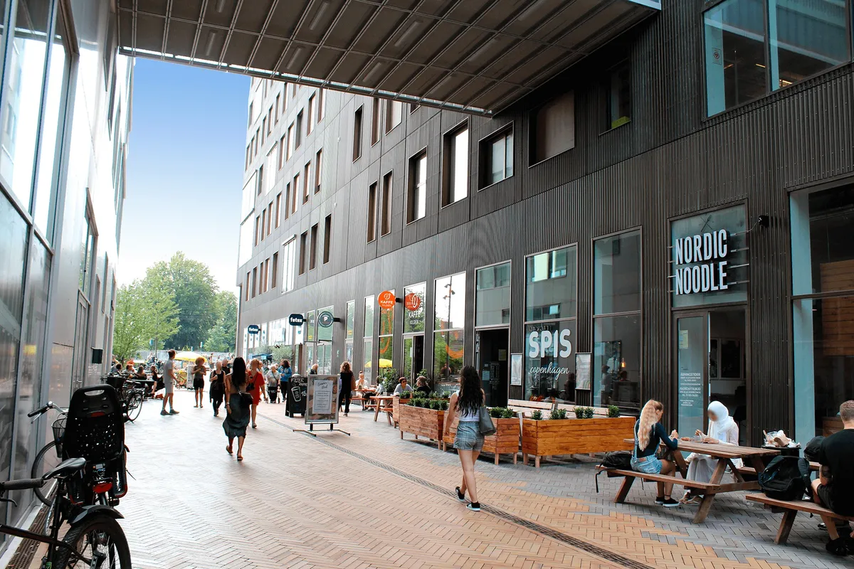 Mennesker går på gågaden i Carlsberg Byen med restauranter og caféer, som Nordic Noodle og Spis Copenhagen, samt træbænke og