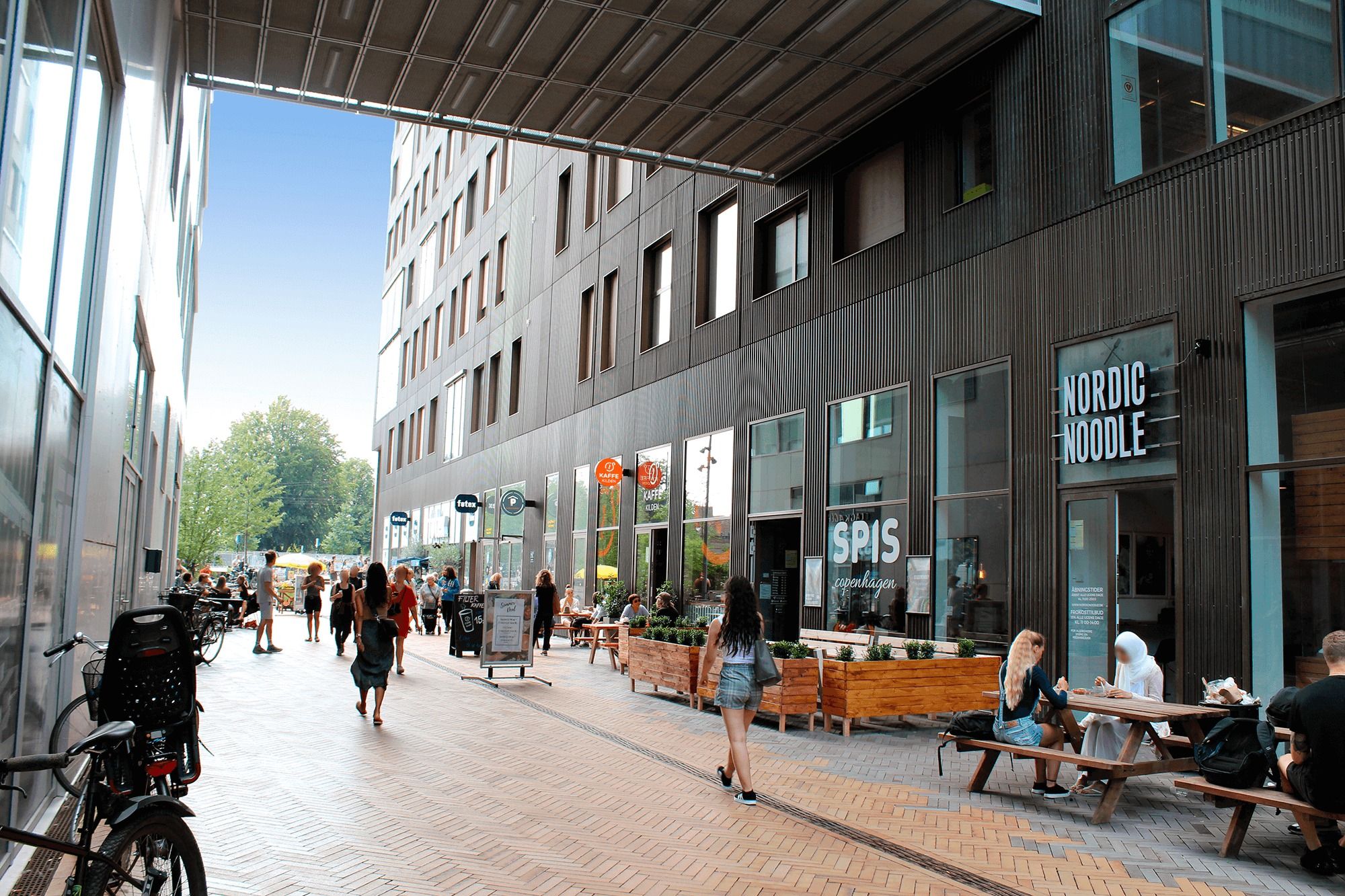 Mennesker går langs gaden og besøger Nordic Noodle, Spis og Kaffe Kolden. Gadeparti med træbænke og planter i potte.