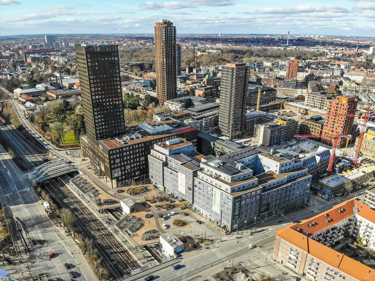 Moderne bygninger med røde tag og flere højhus oprejst ved Carlsberg Byens banegård, omgivet af grønne områder og parker