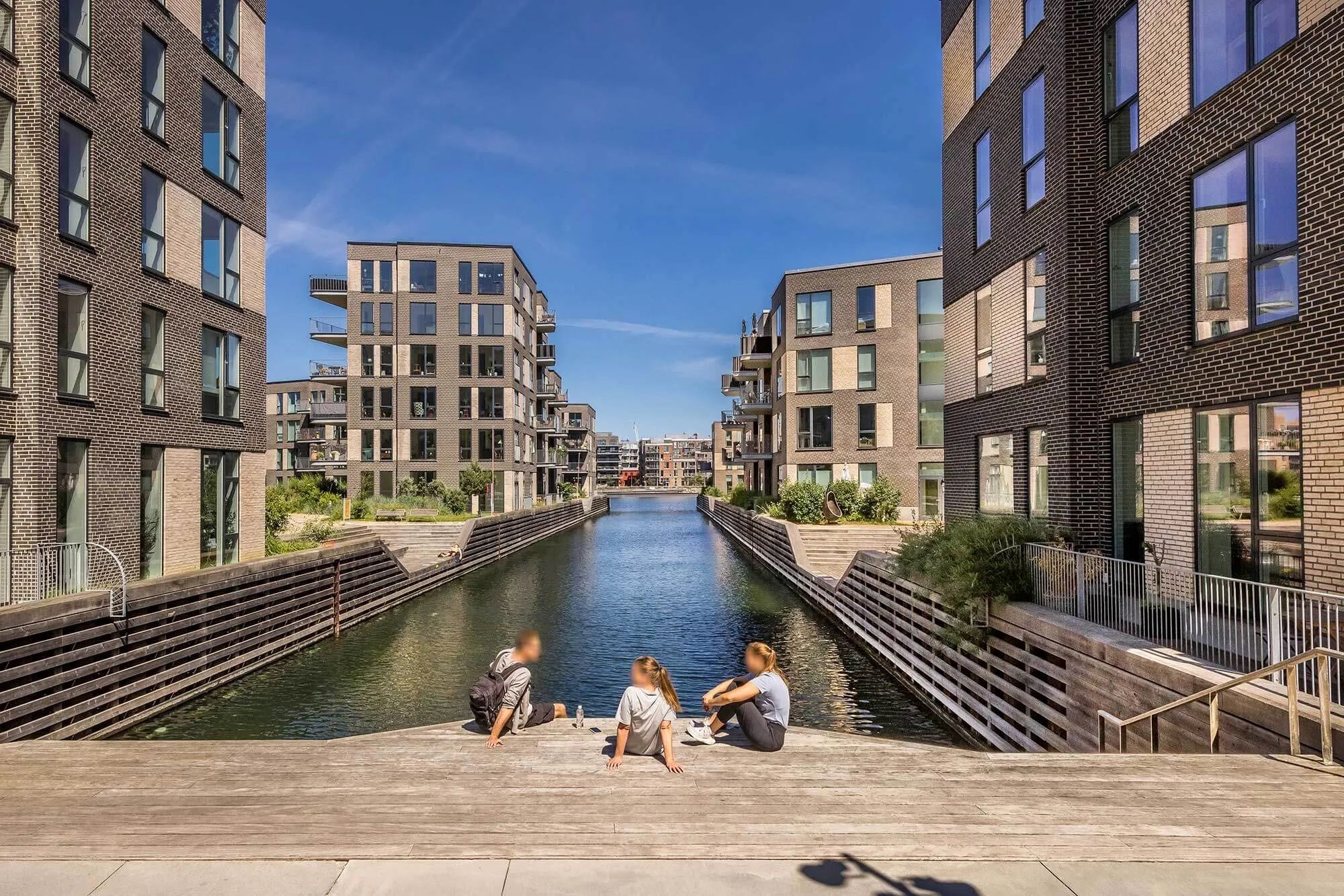Tre unge sidder og slapper af på en træbro over en kanal mellem to højhuse i Nordhavnen