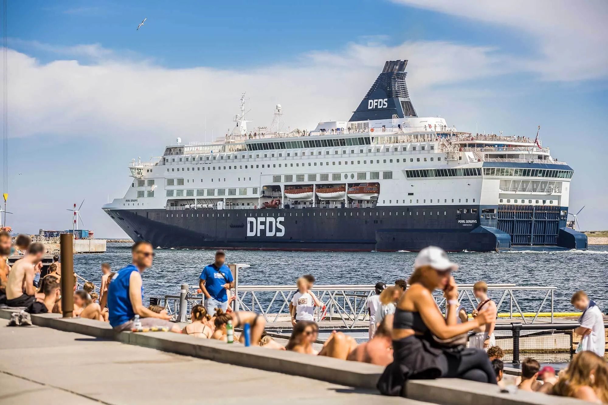 Et stort skib med teksten DFDS ligger ankeret i en havn med solrige og skyggefulde pladser langs kajen
