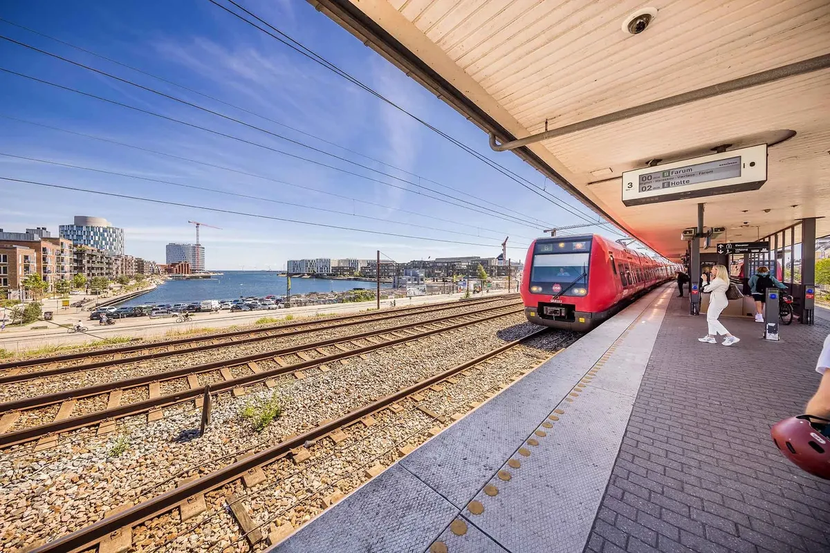 Rød togvogn kører ind på stationen ved havnen, folk venter på perronen og bygninger i baggrunden