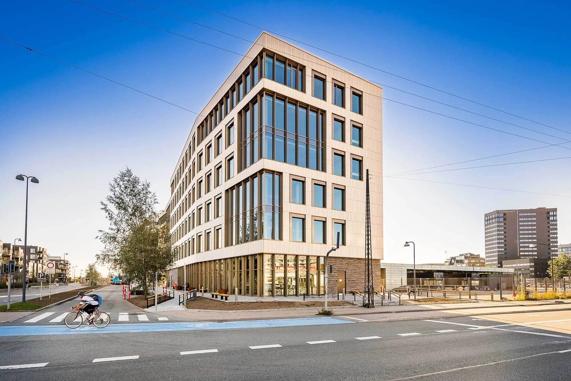 En cykelkører kører på en cykelsti ved en stor bygning med glasvinduer og hjørneplacering i en by