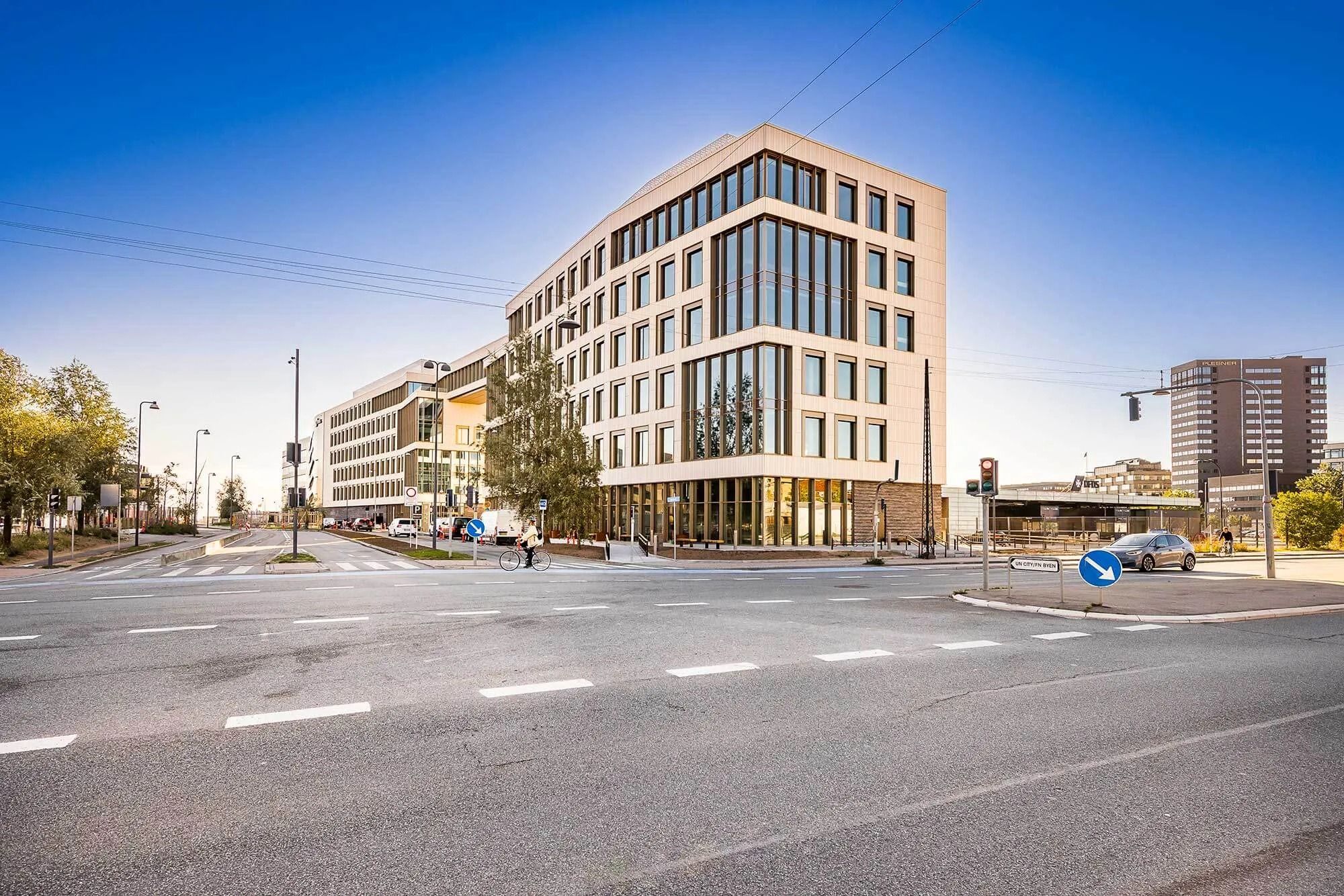 En slank, moderne bygning med store vinduer ligger ved et tomt korsningspunkt med et stort blåt skilt med pile og en