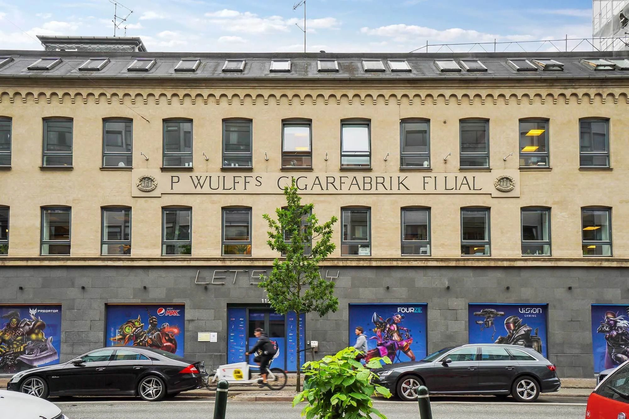 Moderne ejendom med reklame for Pepsi og lejemål med køl. Personer kører på cykler og biler parkeret på gaden.