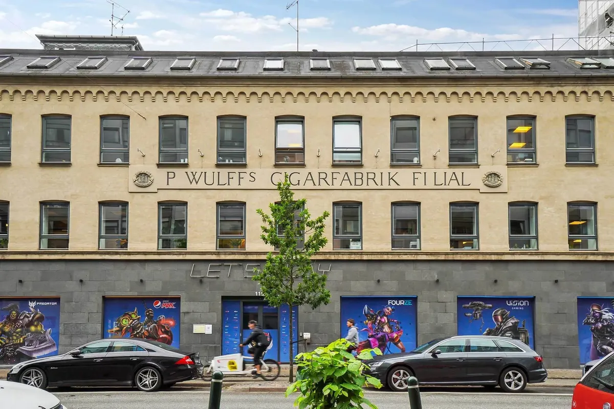 Moderne ejendom med reklame for Pepsi og lejemål med køl. Personer kører på cykler og biler parkeret på gaden.