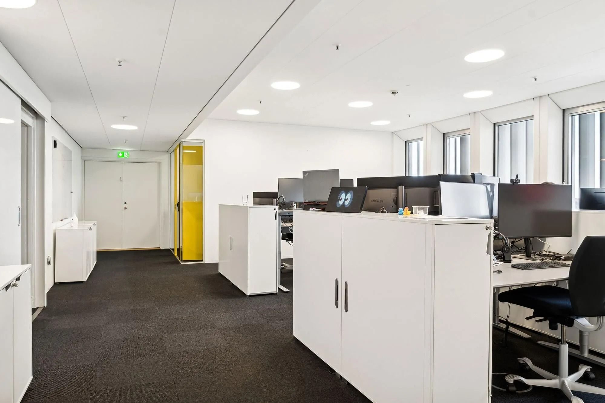 Højt oplyst kontor med hvide vægge, højt hængende runde loftslamper, gul kabinet, hvide skriftborde og computere og gulvtæppe