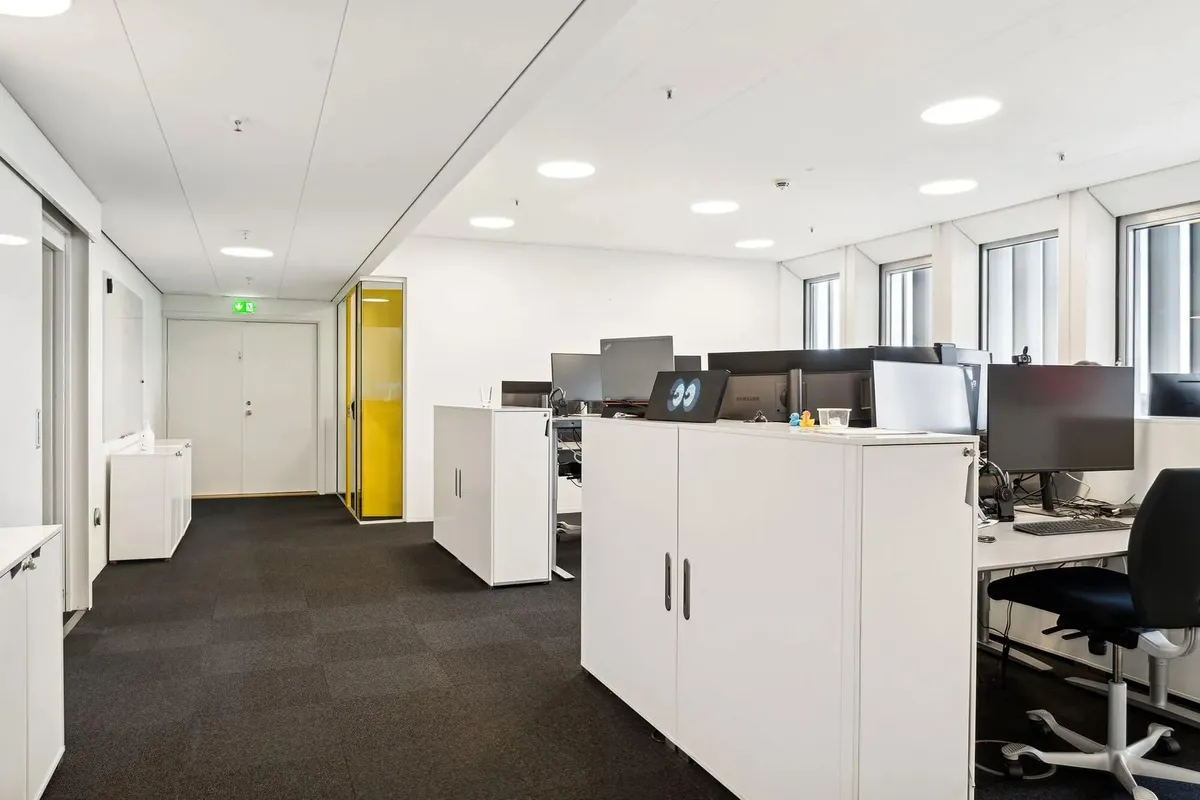 Højt oplyst kontor med hvide vægge, højt hængende runde loftslamper, gul kabinet, hvide skriftborde og computere og gulvtæppe