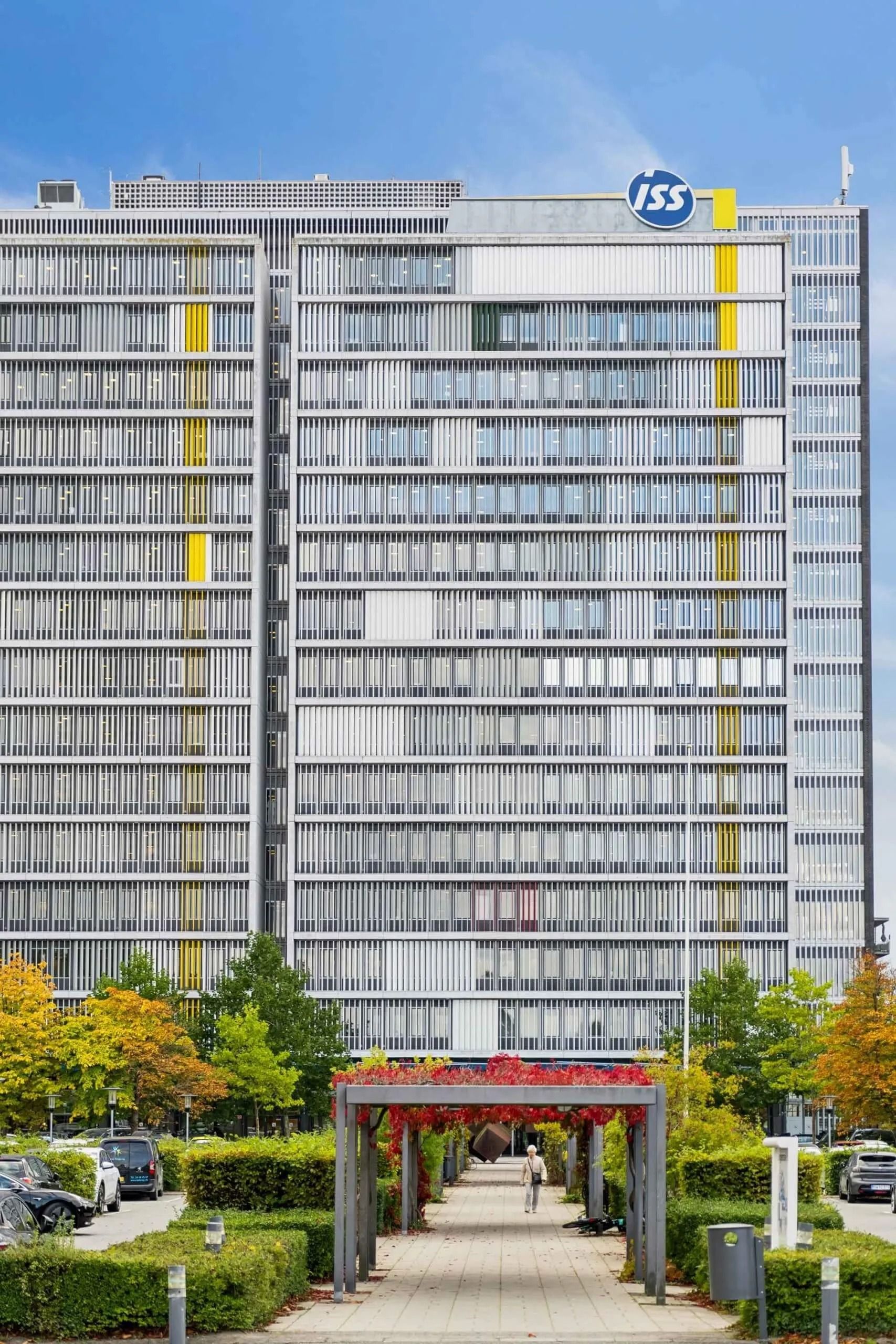 Kontorbygning med moderne design og lyse gule linjer, omgivet af træer og buske, med biler parkeret foran