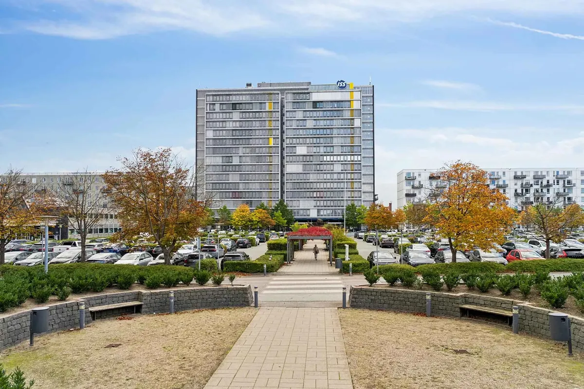 Stort flerbrugskontor med parkeringsplads og vejledningstavle. En person går ad gågaden. Der er flere huse i baggrunden og