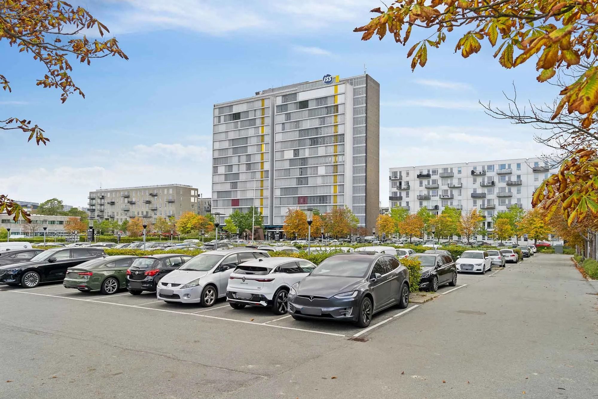 En Tesla Model 3 parkeret i en parkeringsplads med flere biler, med et stort højhus i baggrunden