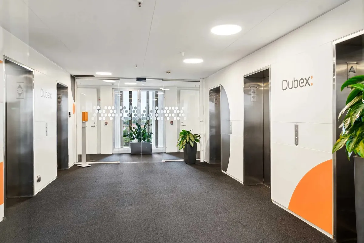 En lys indgangshal med to elevatorer og en dør, der fører til et vinduesområde. Væggene er hvide og der er en stor