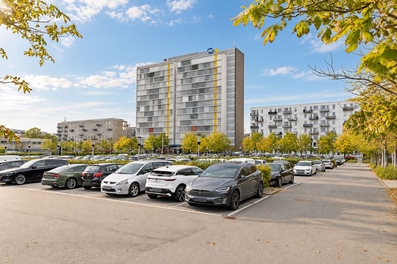 Lyst kontorlejemål i Søborg Tower med parkeringsplads. Forskellige biler parkeret i parkeringsplads. Bygning med gule og hvide striber. Buske og træer i forgrunden.