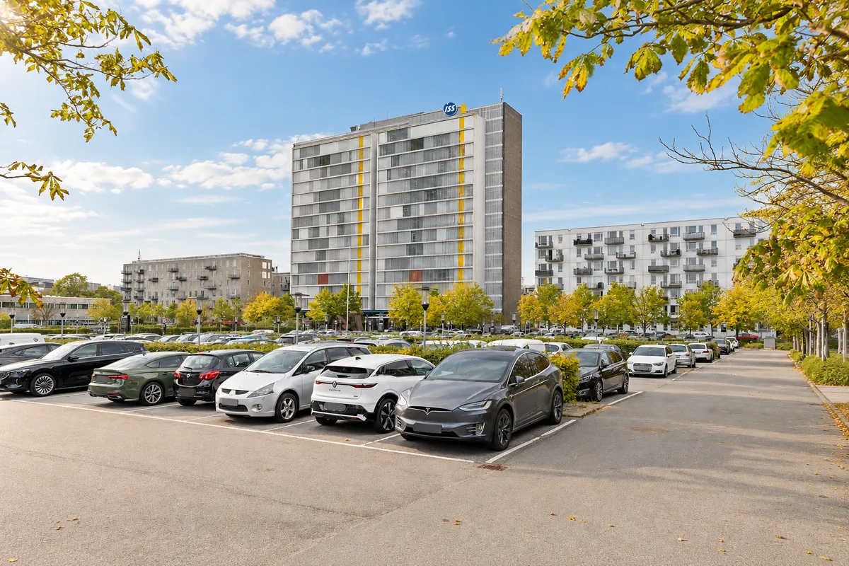 Lyst kontorlejemål i Søborg Tower med parkeringsplads. Forskellige biler parkeret i parkeringsplads. Bygning med gule og hvide striber. Buske og træer i forgrunden.