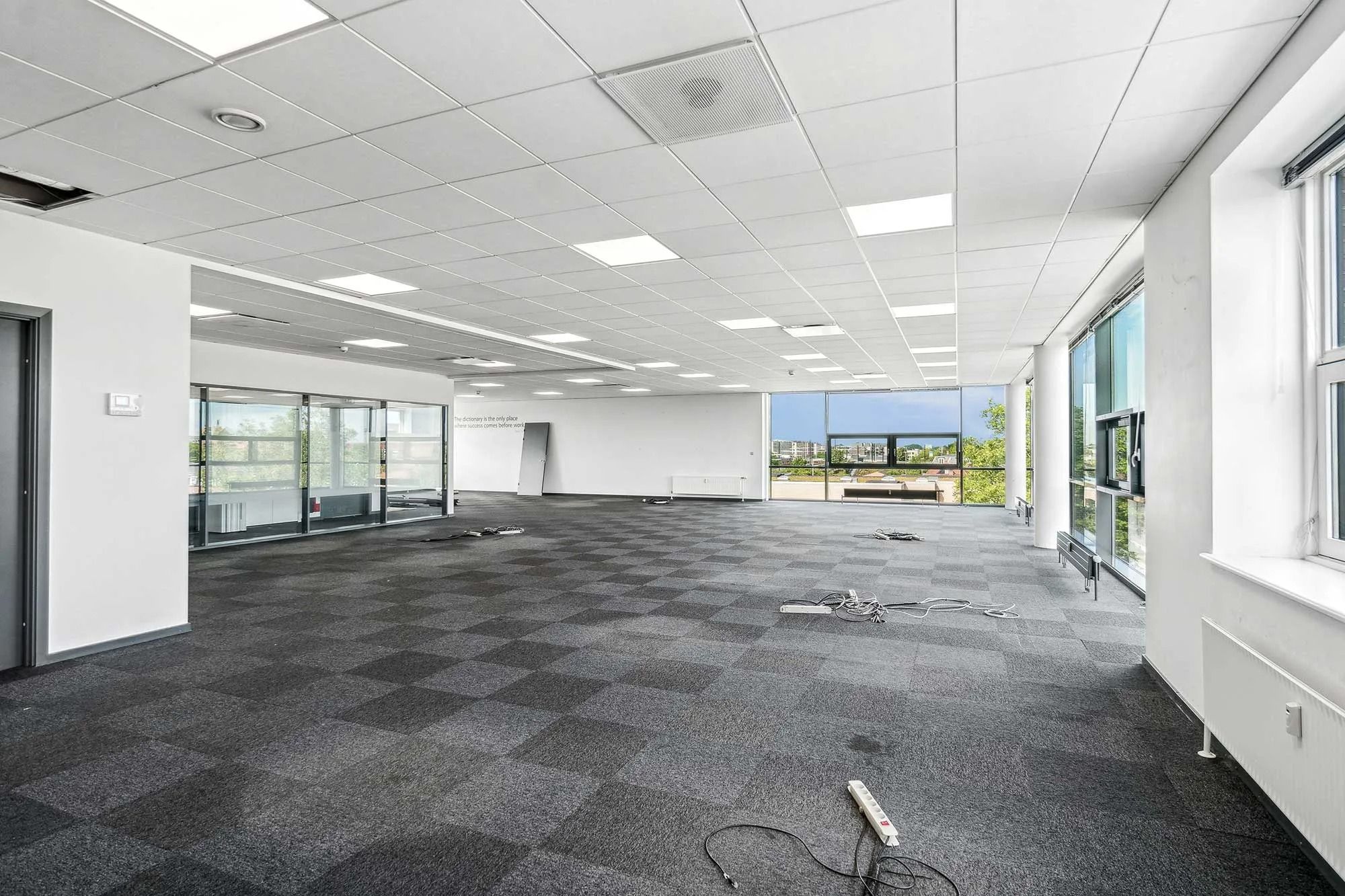 Et tomt kontorrum med en tæppegulv og glasvinduer med udsigt til byen og grønne træer