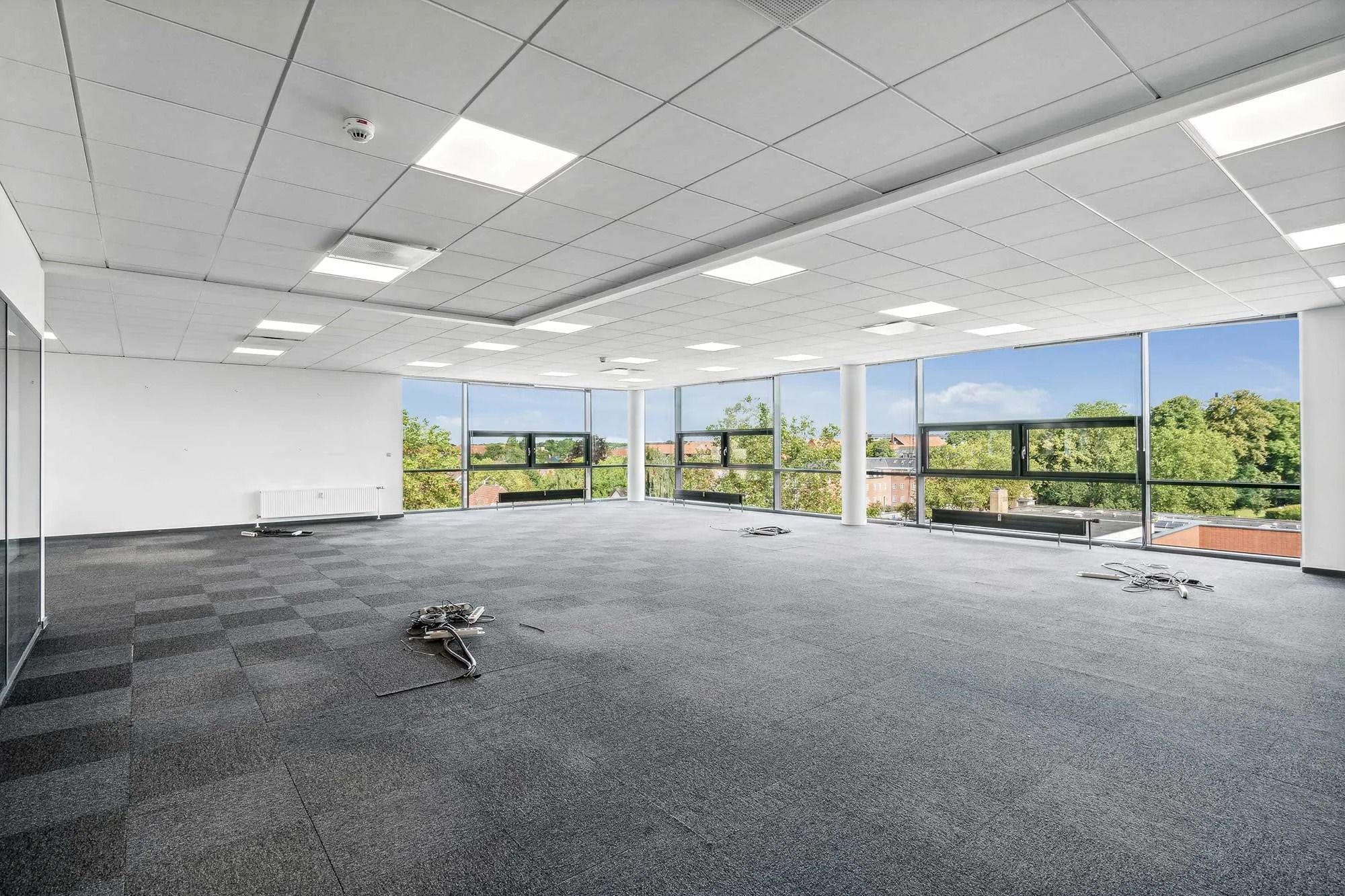 Et stort kontorrum med hvide vægge, gulv og loft samt store vinduer og tæppegulve. Der er en radiator og en sikkerhedskamera