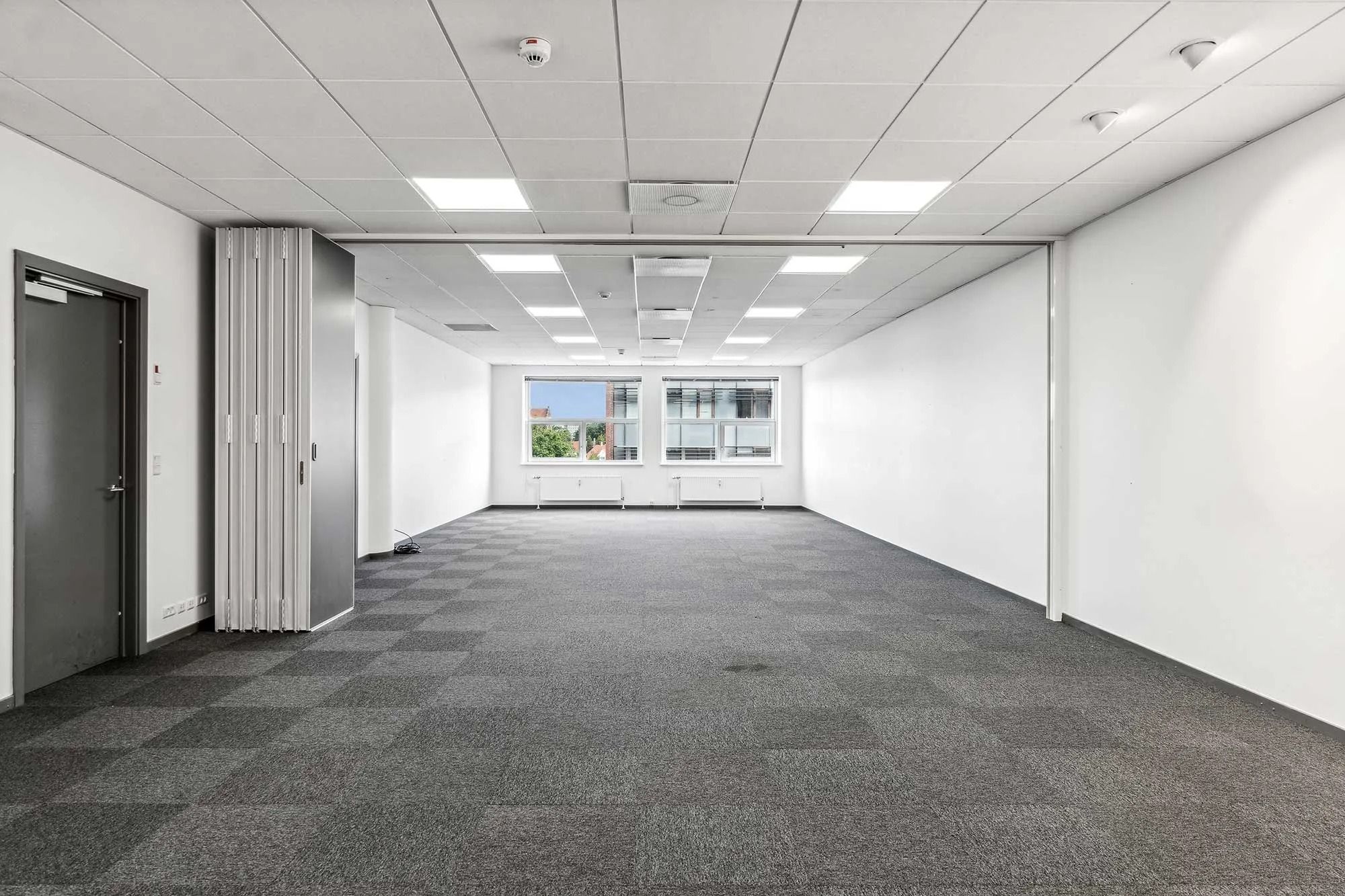 Et stort, ubeboet kontorrum med hvidt loft og hvide vægge samt grå karpetgulv. Værelset har to store vinduer med