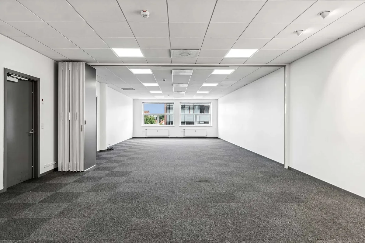 Et stort, ubeboet kontorrum med hvidt loft og hvide vægge samt grå karpetgulv. Værelset har to store vinduer med