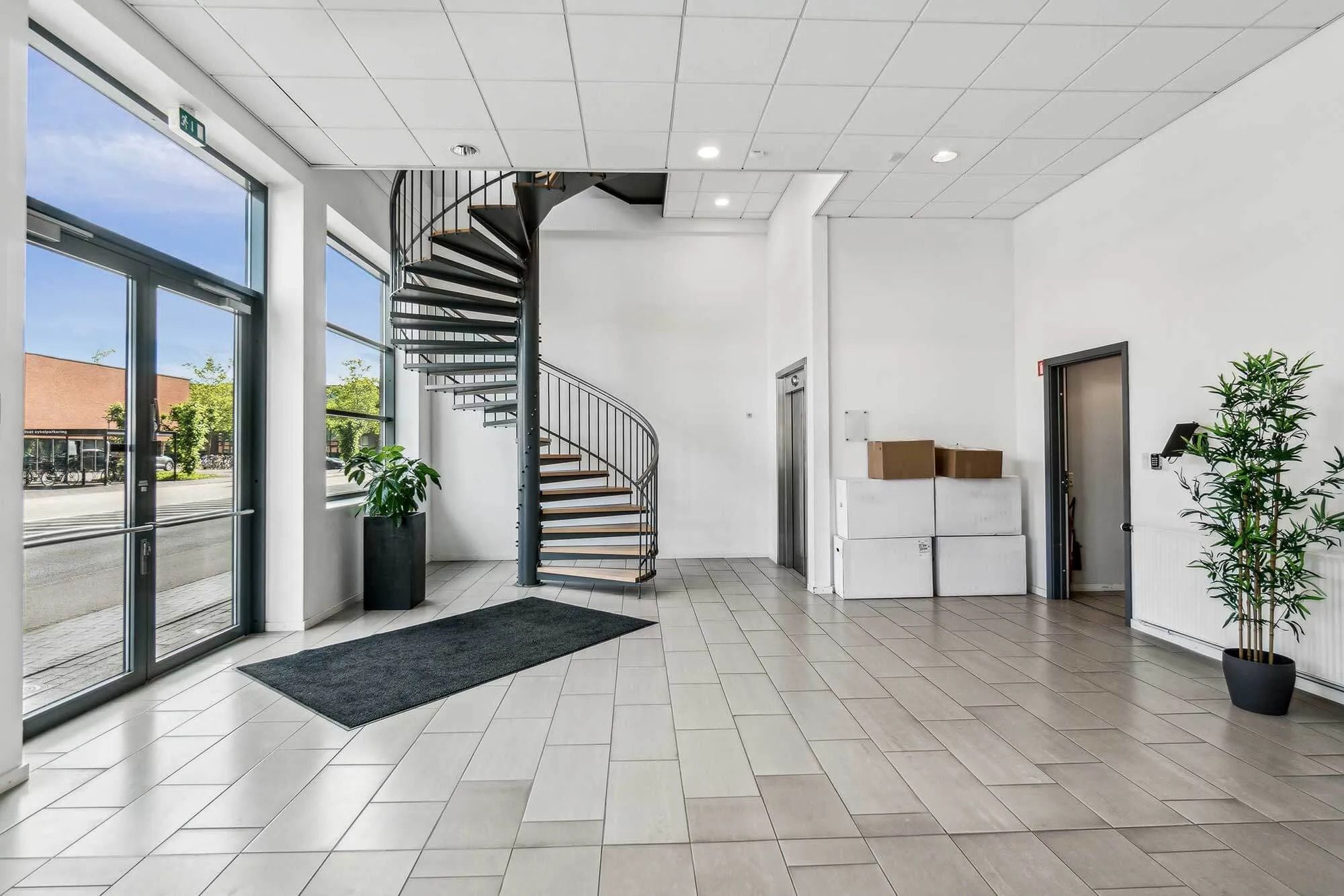 En moderne lobby med spiraltrappe, hvide vægge, højloft, glasdøre og store plastplanter i potter