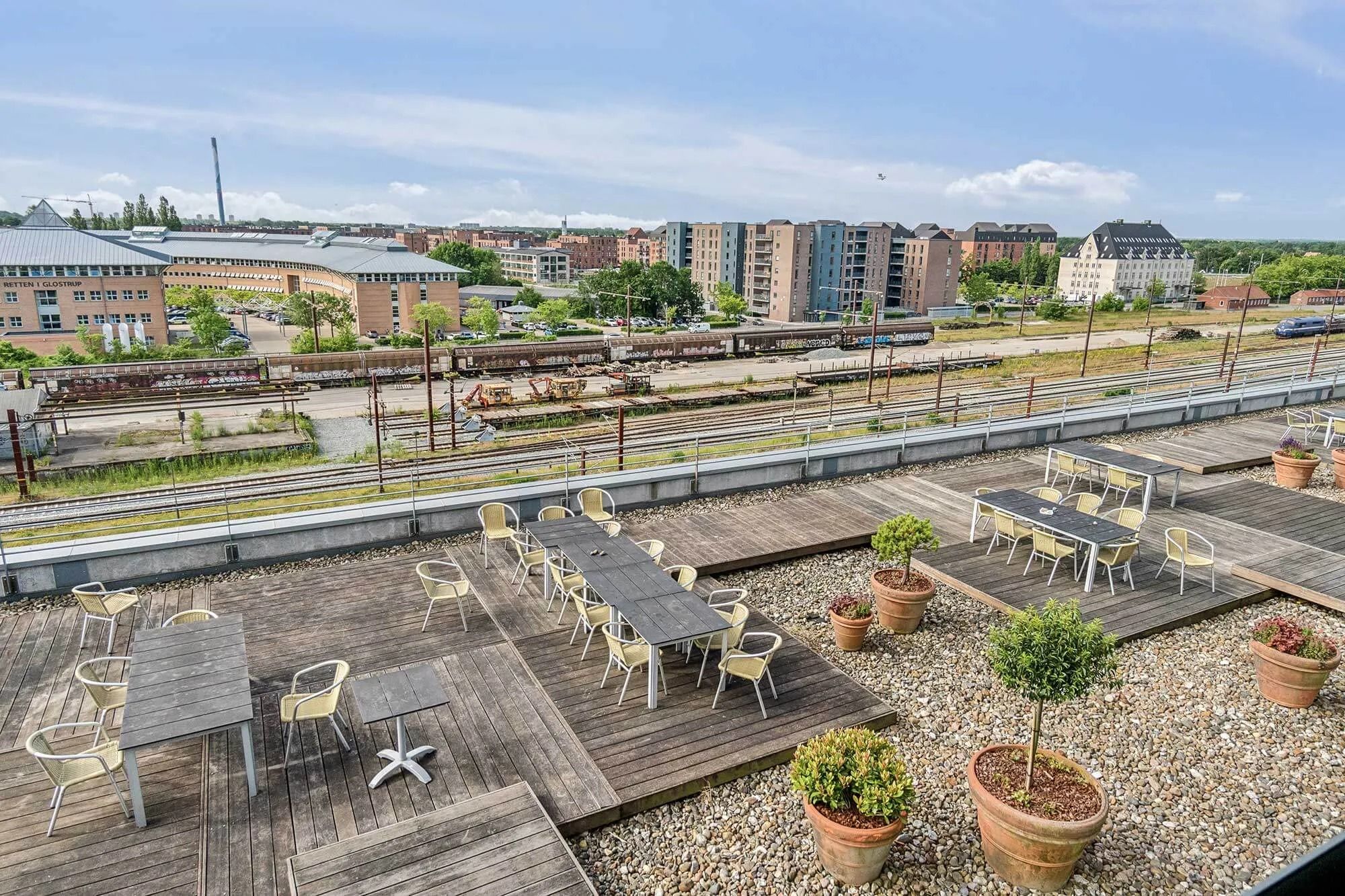 Balkon med flere rekreative møbler i en by. Der er flere træborde og stole samt nogle plantepoteter