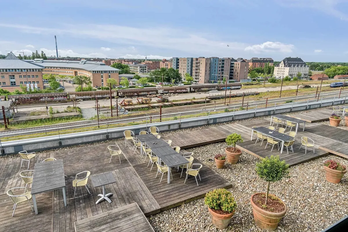 Balkon med flere rekreative møbler i en by. Der er flere træborde og stole samt nogle plantepoteter