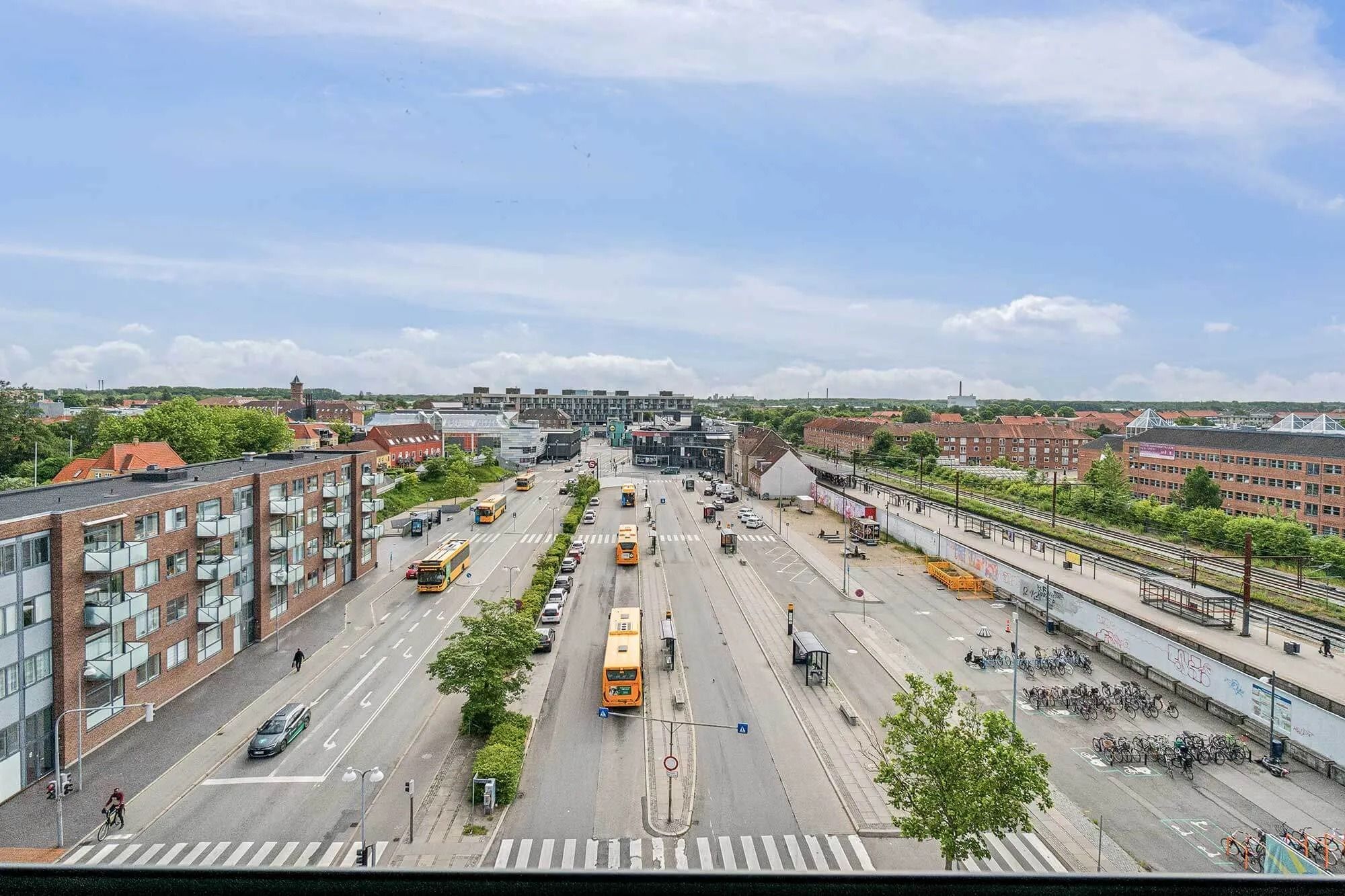 Busser kører gennem en stor vejkrydsning i byen, med grønne busser og flere biler. På den ene side af vejen er der