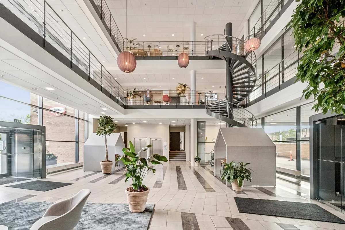Planter og moderne møbler sammen med lyse loftslampe og glasdøre i kontorlobbyen med lysegule gulvfliser og mørke grå tæpper