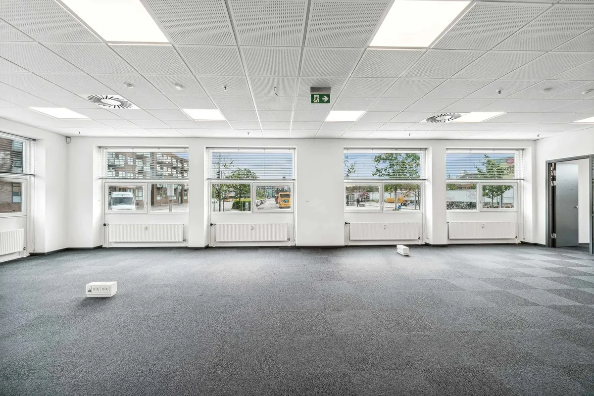 Store kontorlokaler med lyse lofter og hvide vægge. Der er fire store ruder med jalousier. På gulvet ligger to hvide bokse