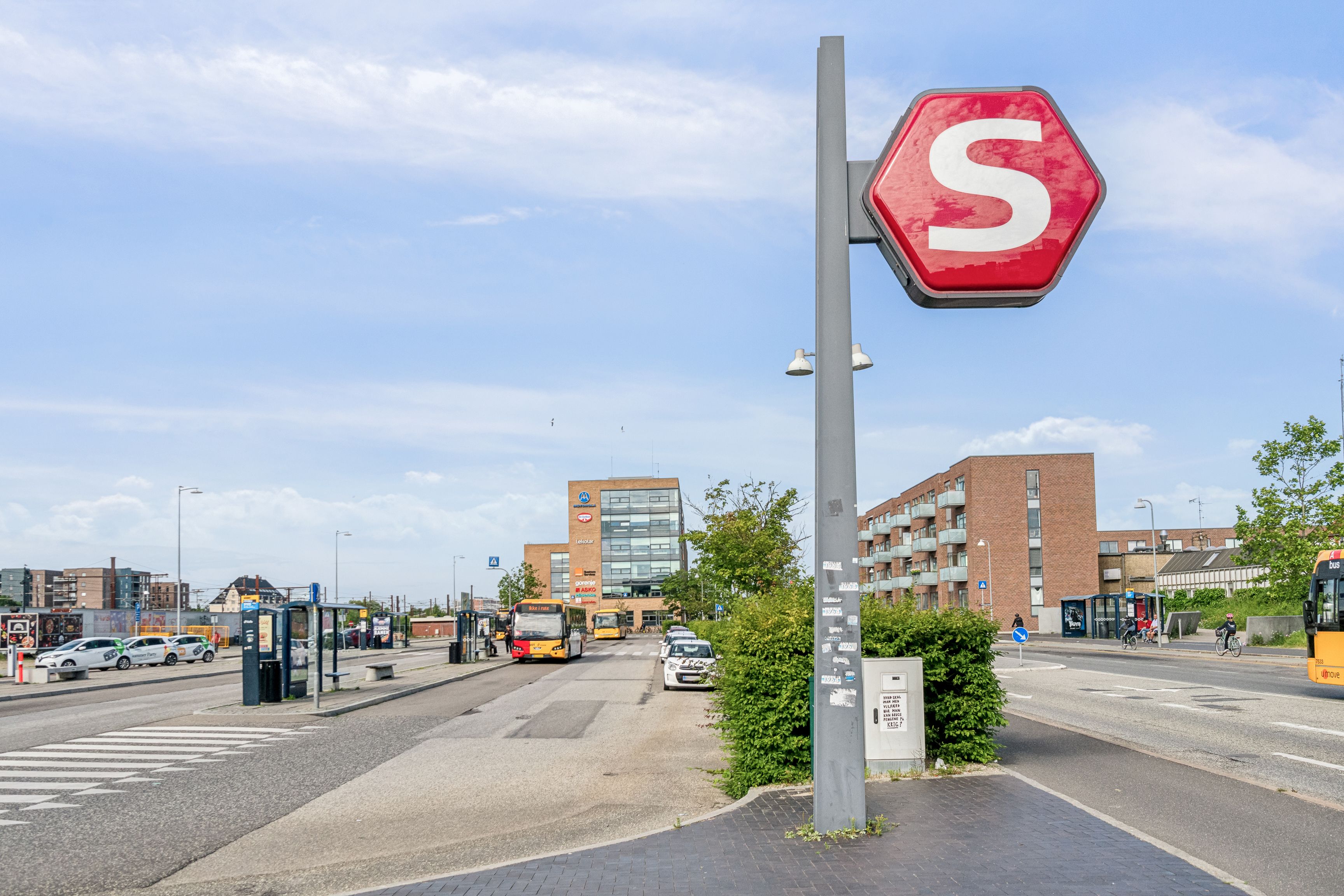 Strømforsyningssøjle med stopskilt og videoovervågning nær busstoppested med biler og cyklister på en travl vej med bygninger og træer i baggrunden