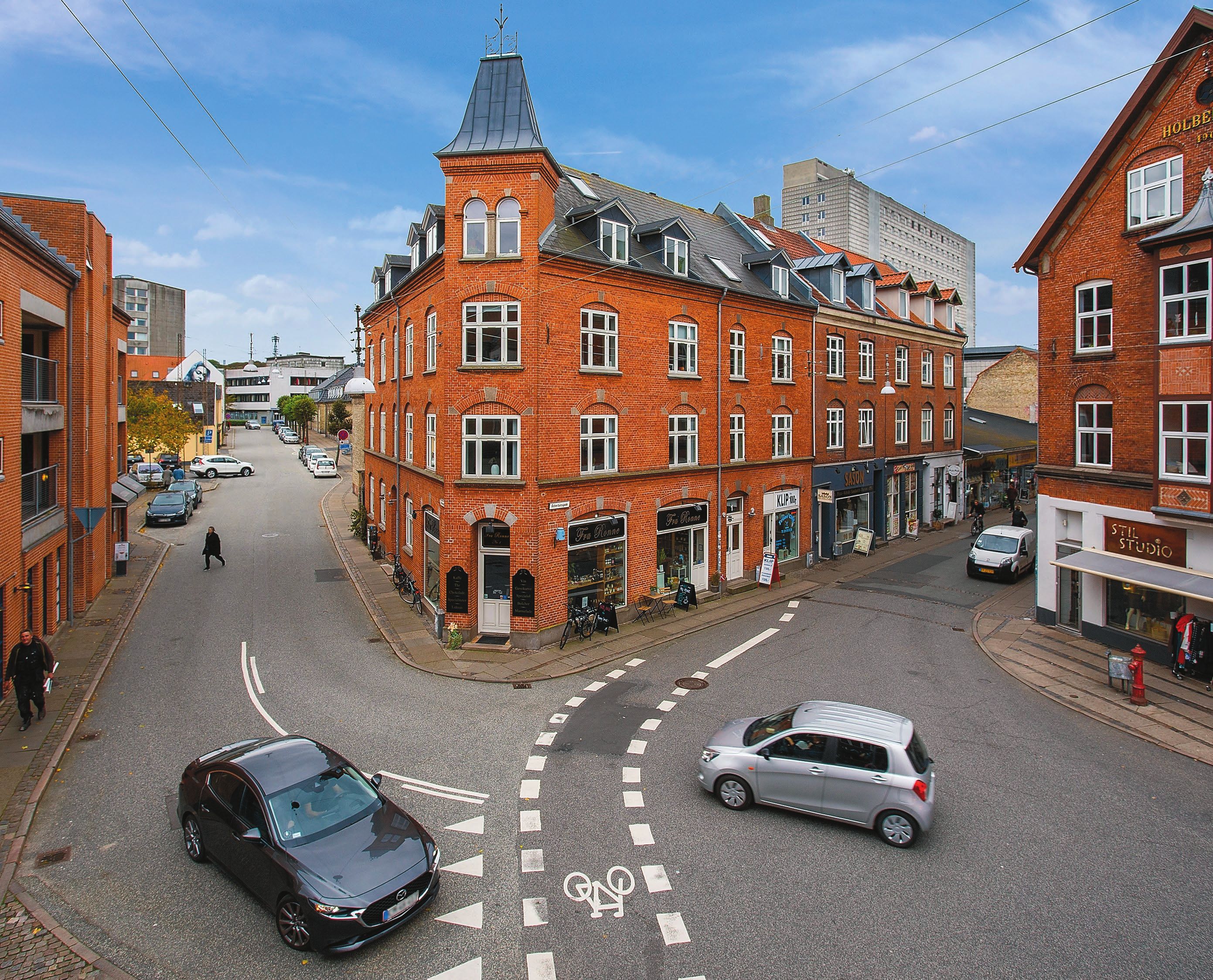 To biler kører rundt på et rundkørsel og en person går på fodgængerfeltet, foran en butik med hyggelig kaffestue