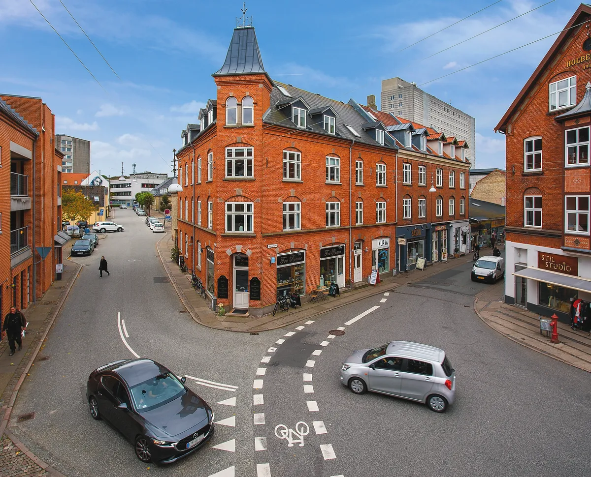 To biler kører rundt på et rundkørsel og en person går på fodgængerfeltet, foran en butik med hyggelig kaffestue