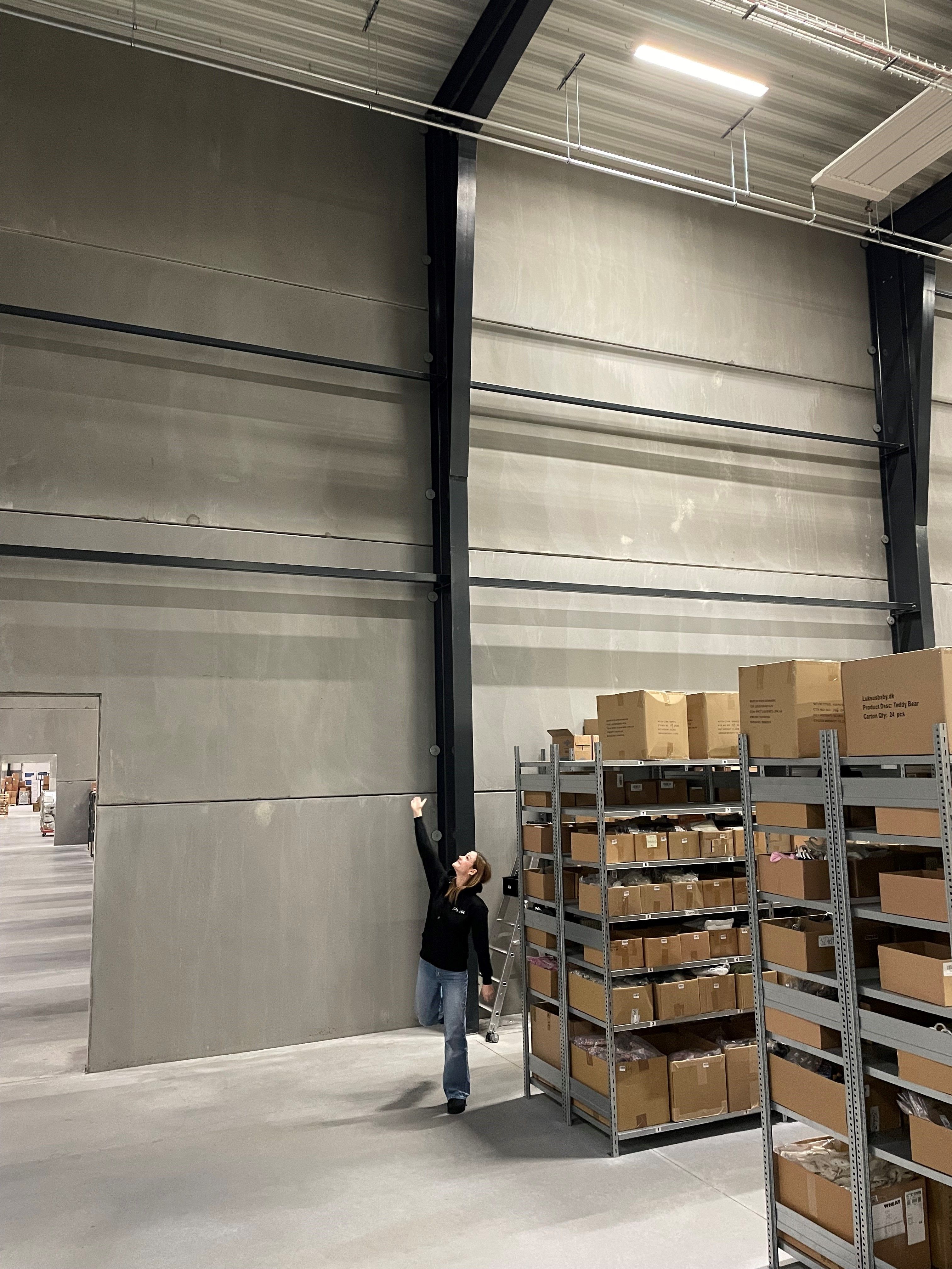 En person står og strækker armen mod loftet i et lager med flere reoler fyldt med kasser med produkter