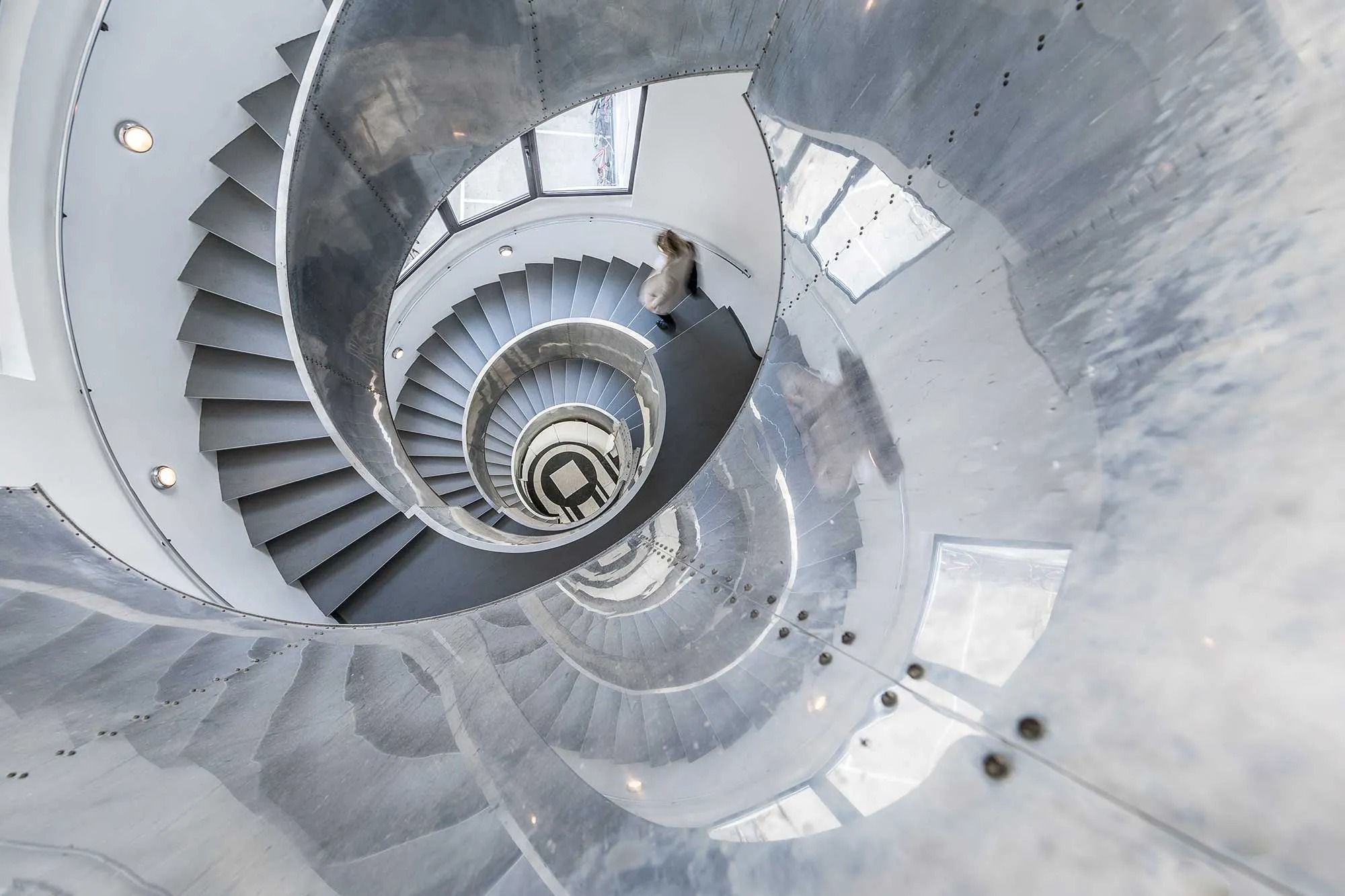 Spiraltrapper med metal og glas, der reflekterer og viser detaljer og mønstre, samt en person, der går i spiralen