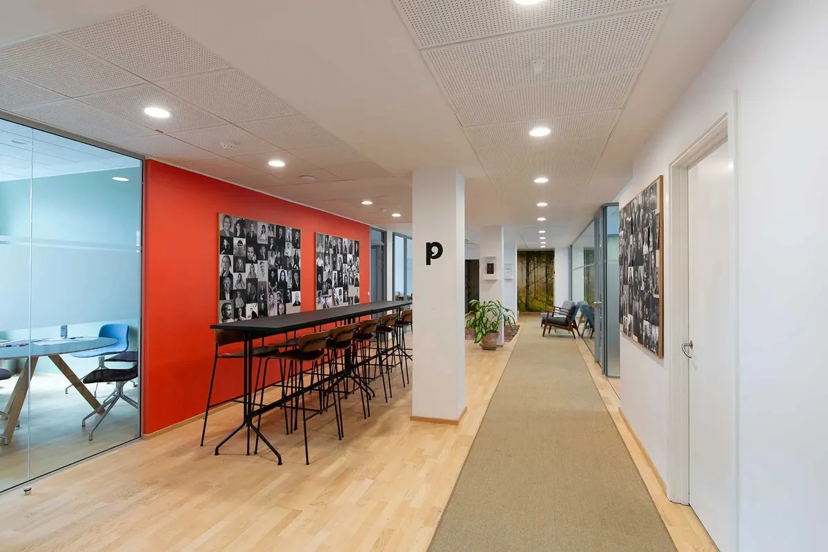 Kontorgang med fotokollage og mødebord på glas og træ, med gulvvæg og loftslamper
