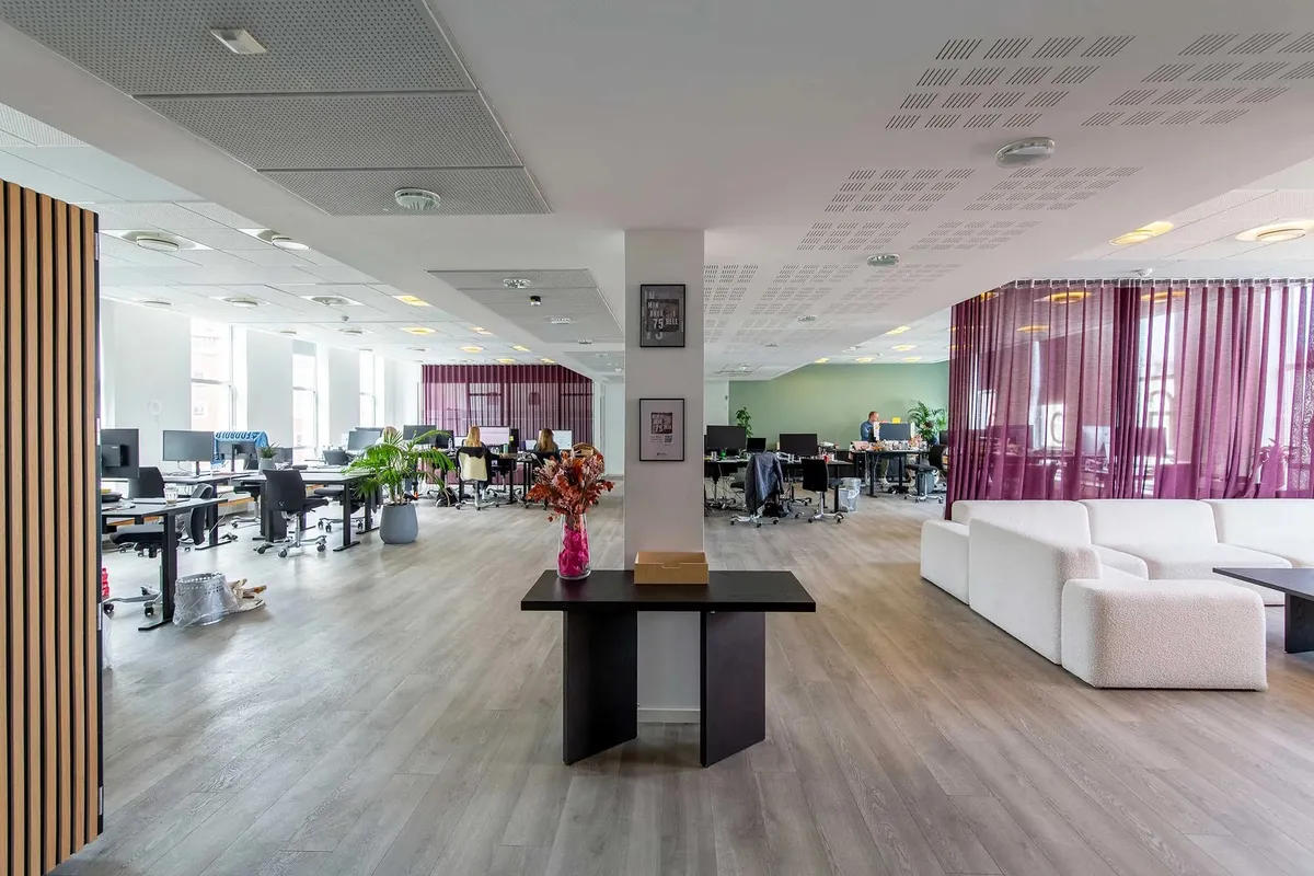 Moderne og åben kontor med højt loft, lysegult gulv og bord med vase, hvor flere mennesker sidder og arbejder