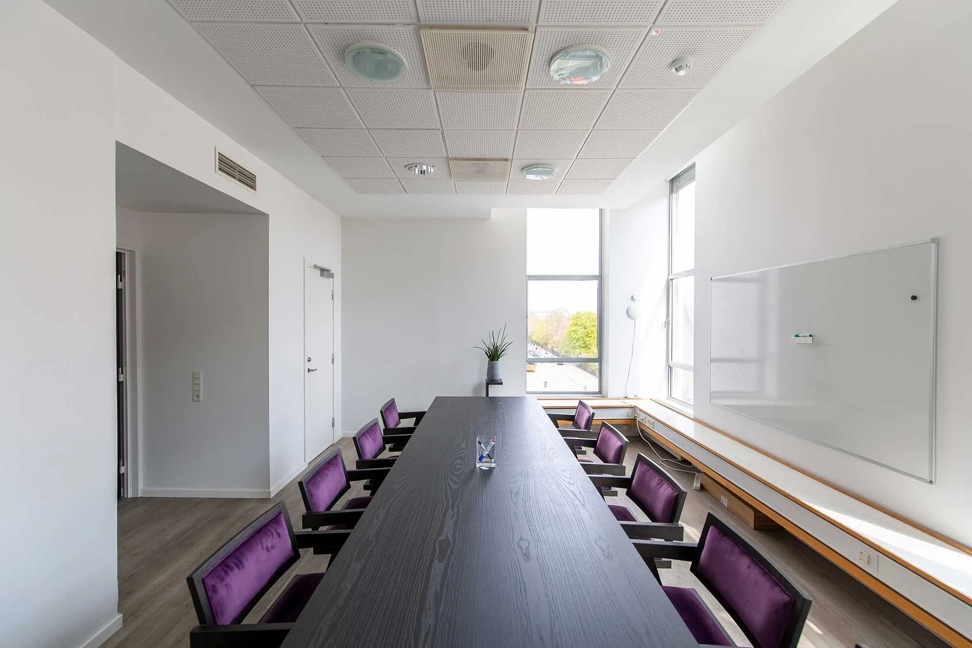 En mødelokale med højt loft, hvide vægge og lysegult gulv samt borde og stole i mørkt farvede stoffer og en stor whiteboard