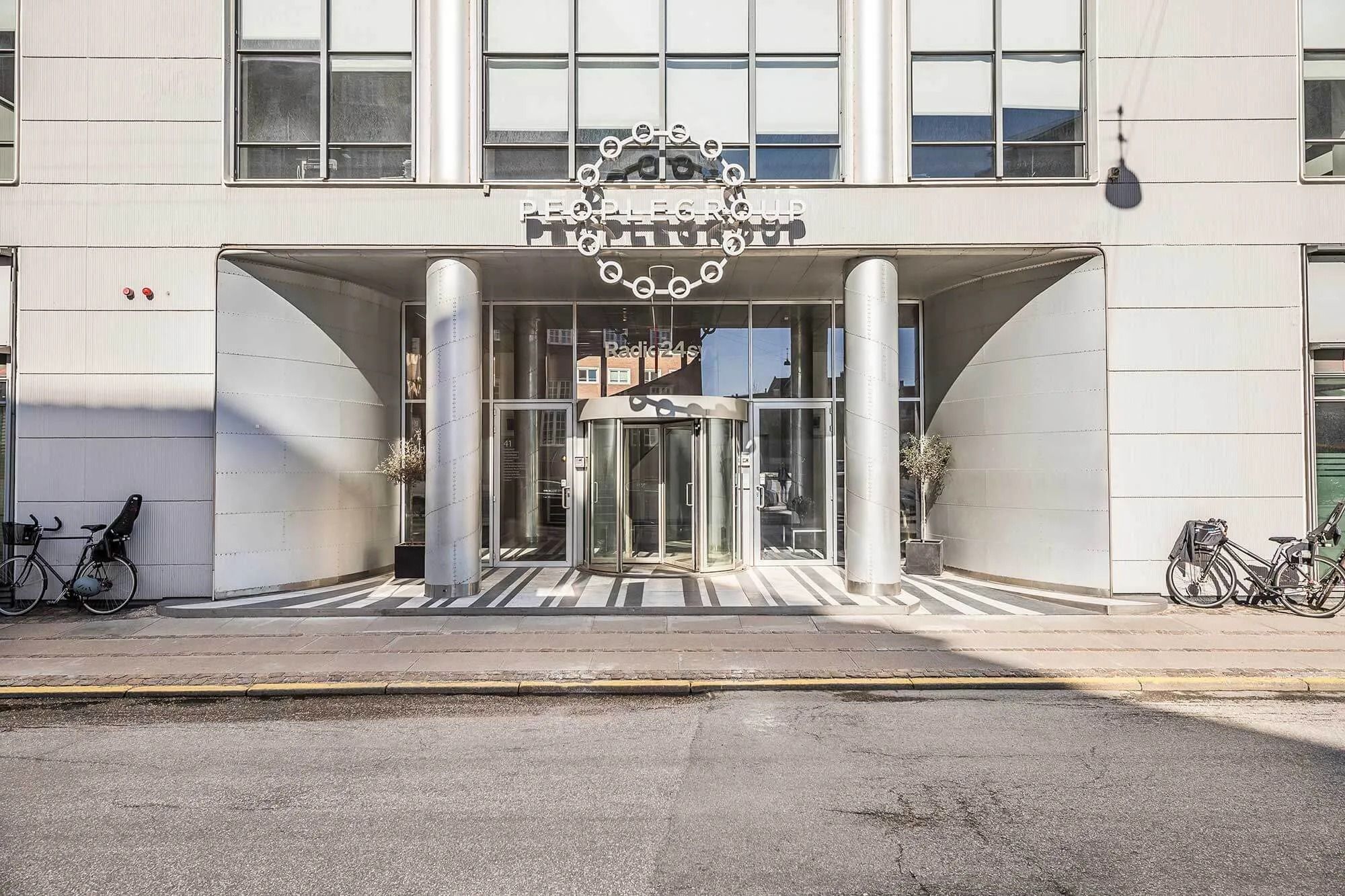 Fremtrædende entre til People Group med søjler og rund logo i hoveddøren med glasskærme og to planter til hver side