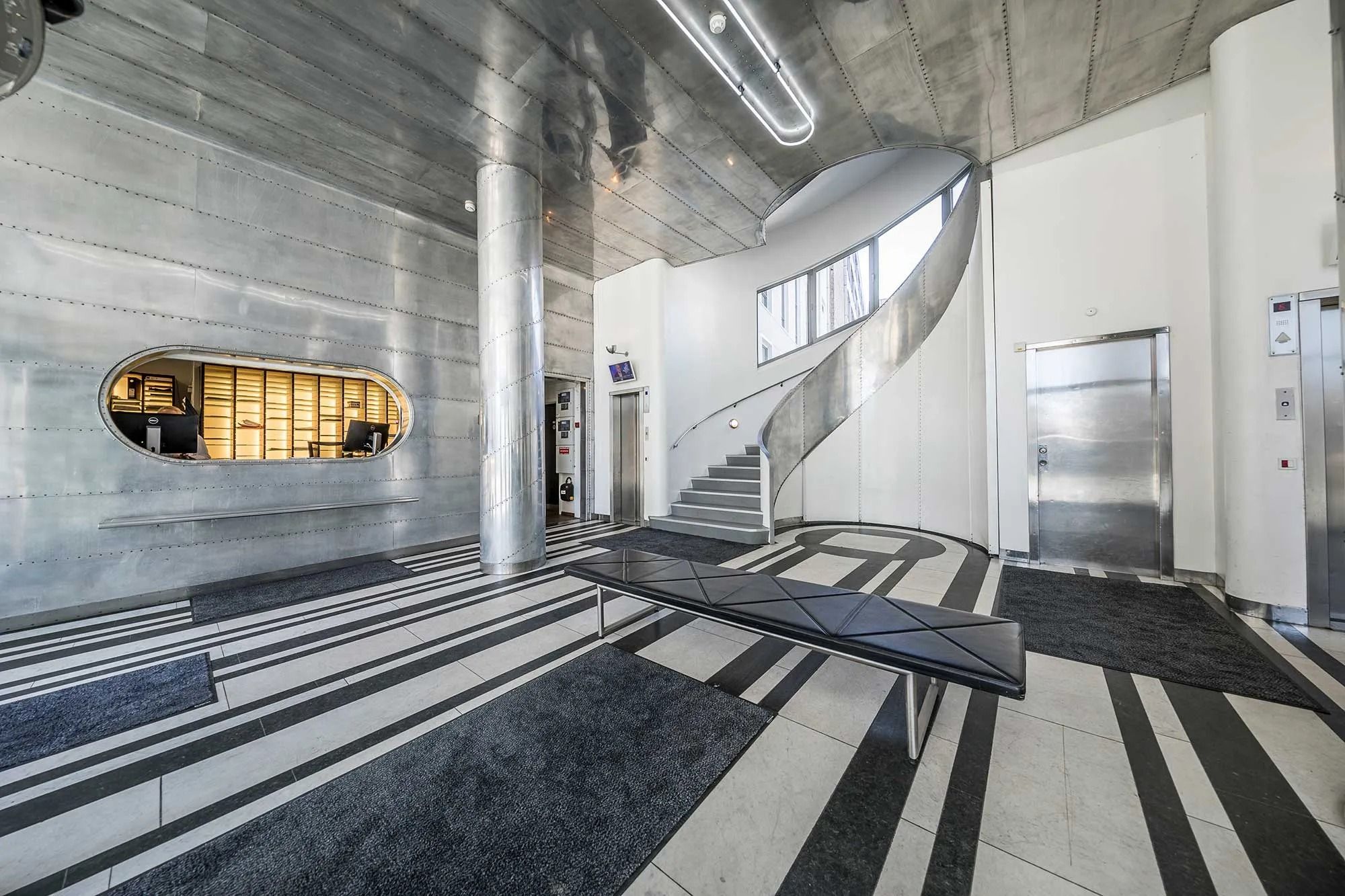 Moderne lobby med lyse metalvægge og skrå stige, en slank sofabank og flisegulv med tydelige farvede striber