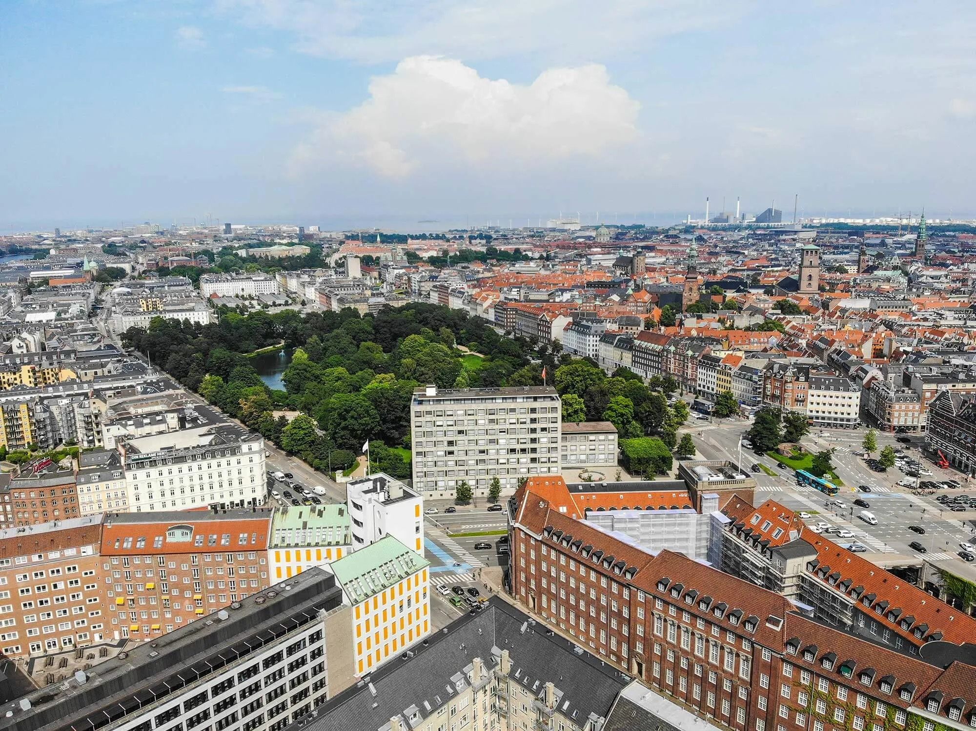 Billede af byen København med park, bygninger, veje, biler, træer og busser i lyseregn på en klar dag