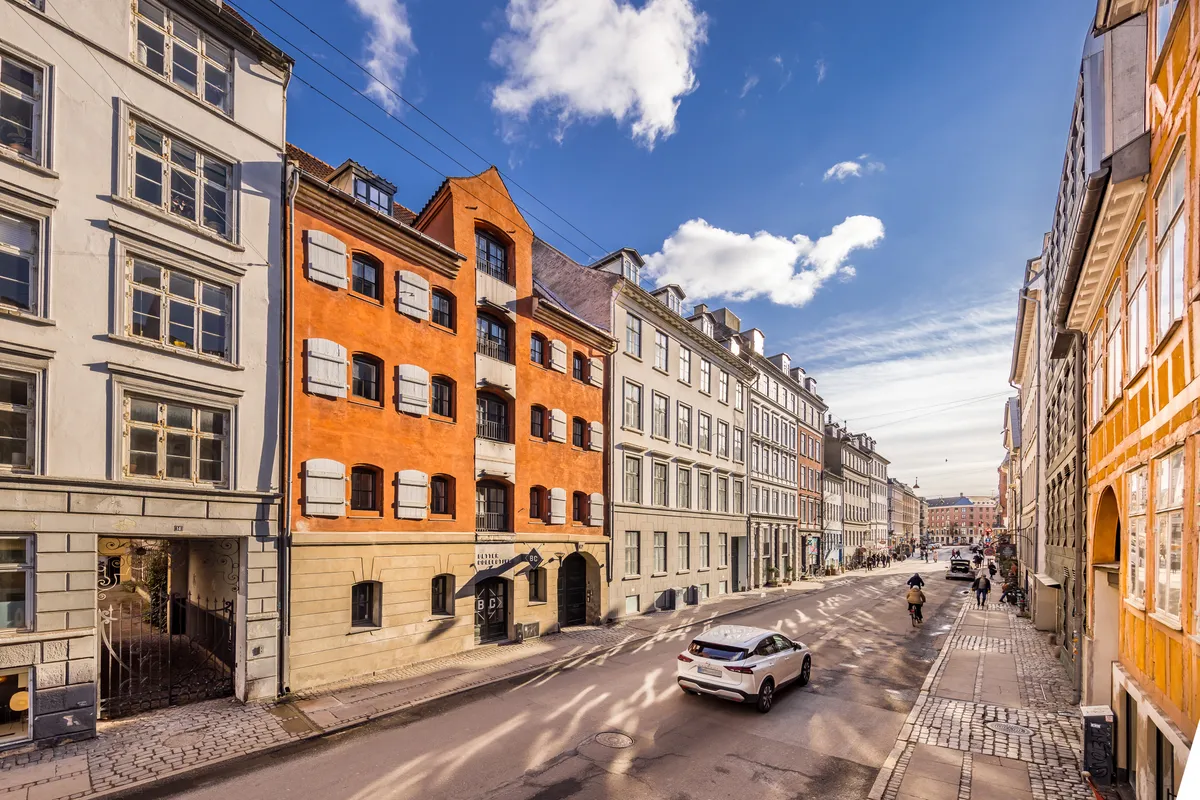 En bil kører langs en brostensbelagt gade i København med mange historiske bygninger, og folk går på fortovet