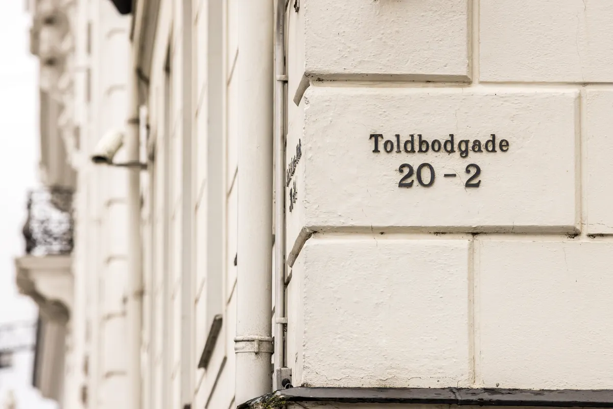 En hvid husvæg med en skilt, der læser "Tolbødgade 20-2" og et overvågningskamera på gulvejret