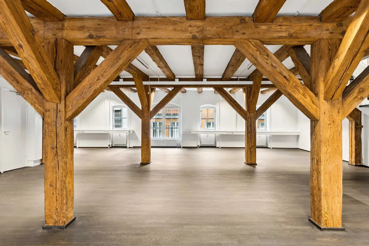 Store bjælker og loftskonstruktioner i en stor stue. Trægulv og lyse vægge. Stående radiatorer og vinduer med udsigt til