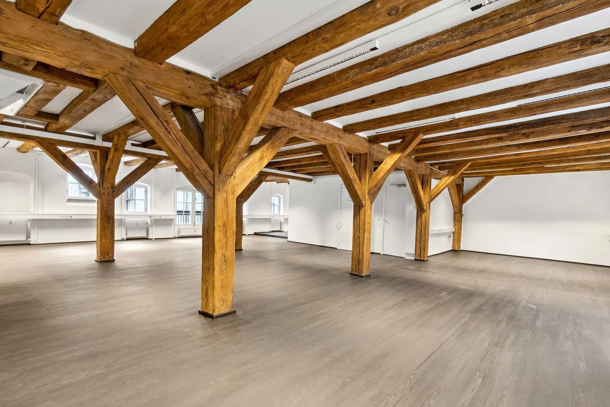 En stor indre rum med lyse vægge og træbjælker. Jorden er dækket med lysegrå trægulv og der er hvide loftsbjælker.