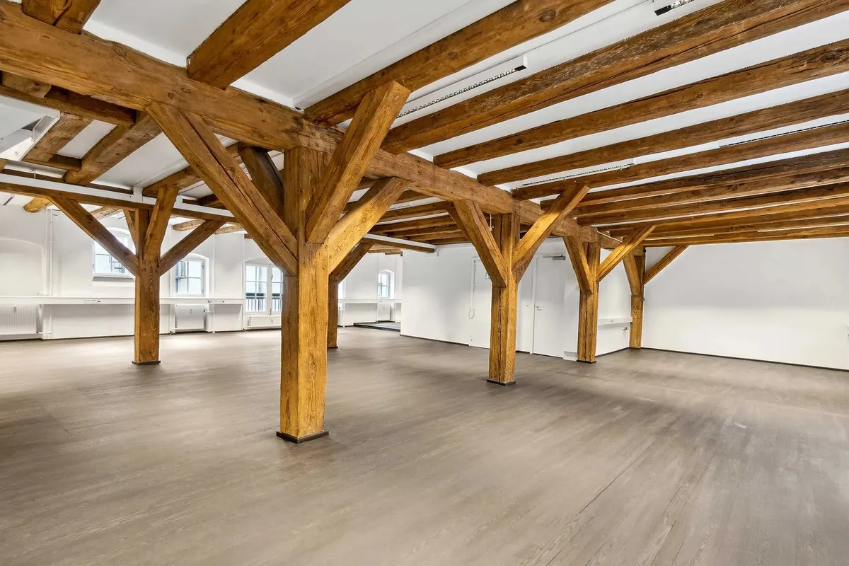 En stor indre rum med lyse vægge og træbjælker. Jorden er dækket med lysegrå trægulv og der er hvide loftsbjælker.